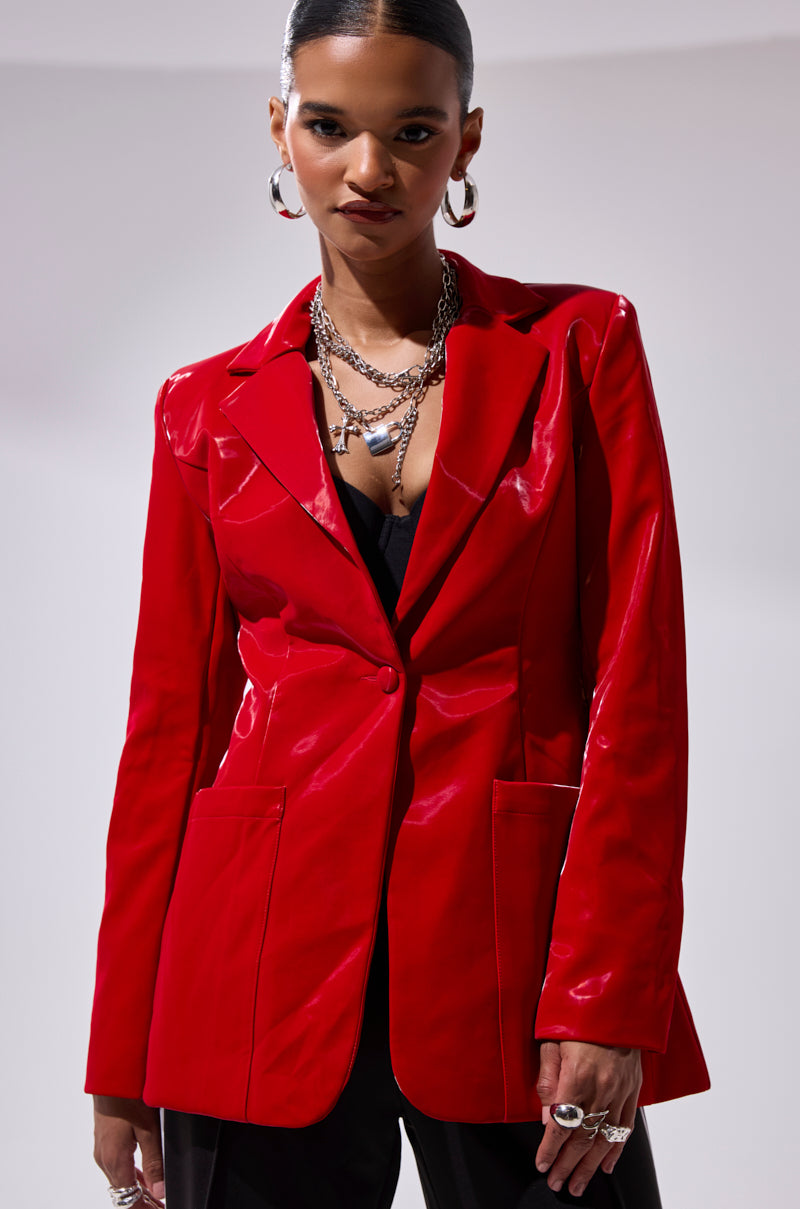 A woman stands confidently against a plain light background in the TOO SLICK LIQUID VELVET BLAZER, a shiny red faux patent leather piece styled over a black top with layered silver necklaces, hoop earrings, and rings.