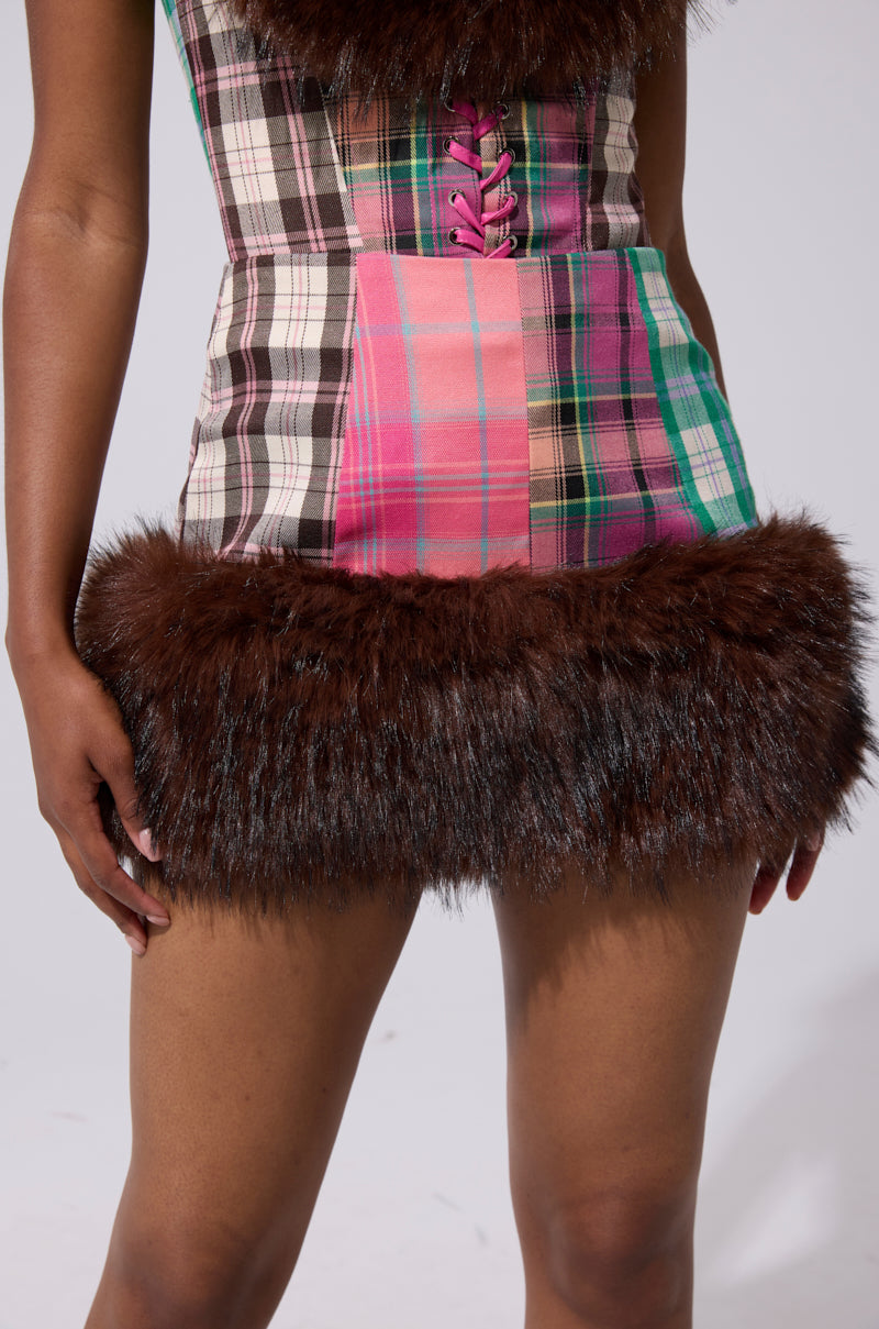 Wearing the SWEET THING PATCHWORK MINI SKIRT and matching top with pink front lacing and brown faux fur trim on the neckline and hem, a person stands against a plain background.