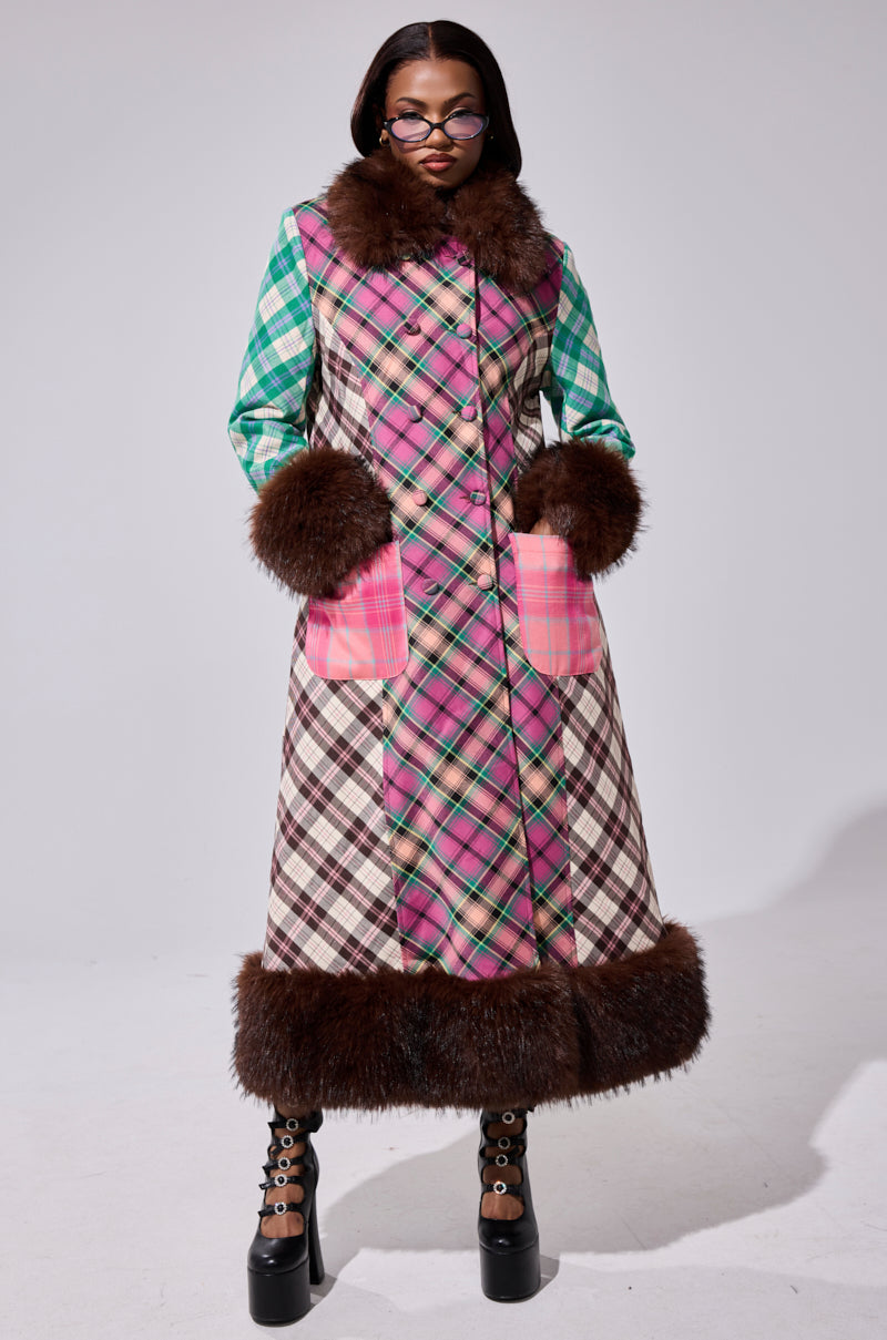 A woman stands confidently in the SWEET THING PATCHWORK TRENCH, a pink plaid coat with faux fur trim on the collar, cuffs, and hem. She wears glasses, platform boots, and has her hands in her pockets.