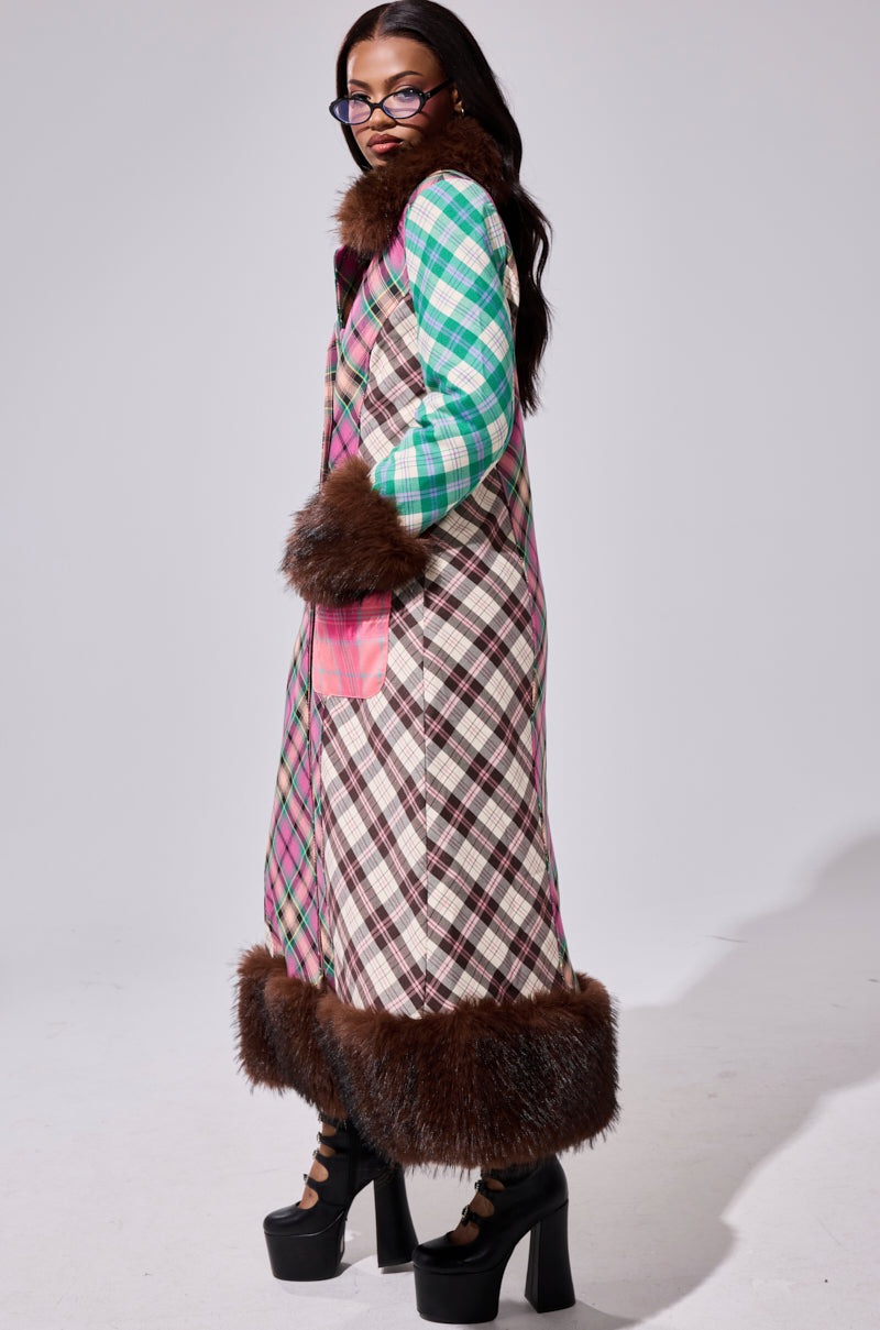 A woman in glasses models the SWEET THING PATCHWORK TRENCH, featuring pink plaid, green checkered sleeves, and faux fur trim. She faces slightly sideways over her shoulder against a plain backdrop, wearing platform heels.