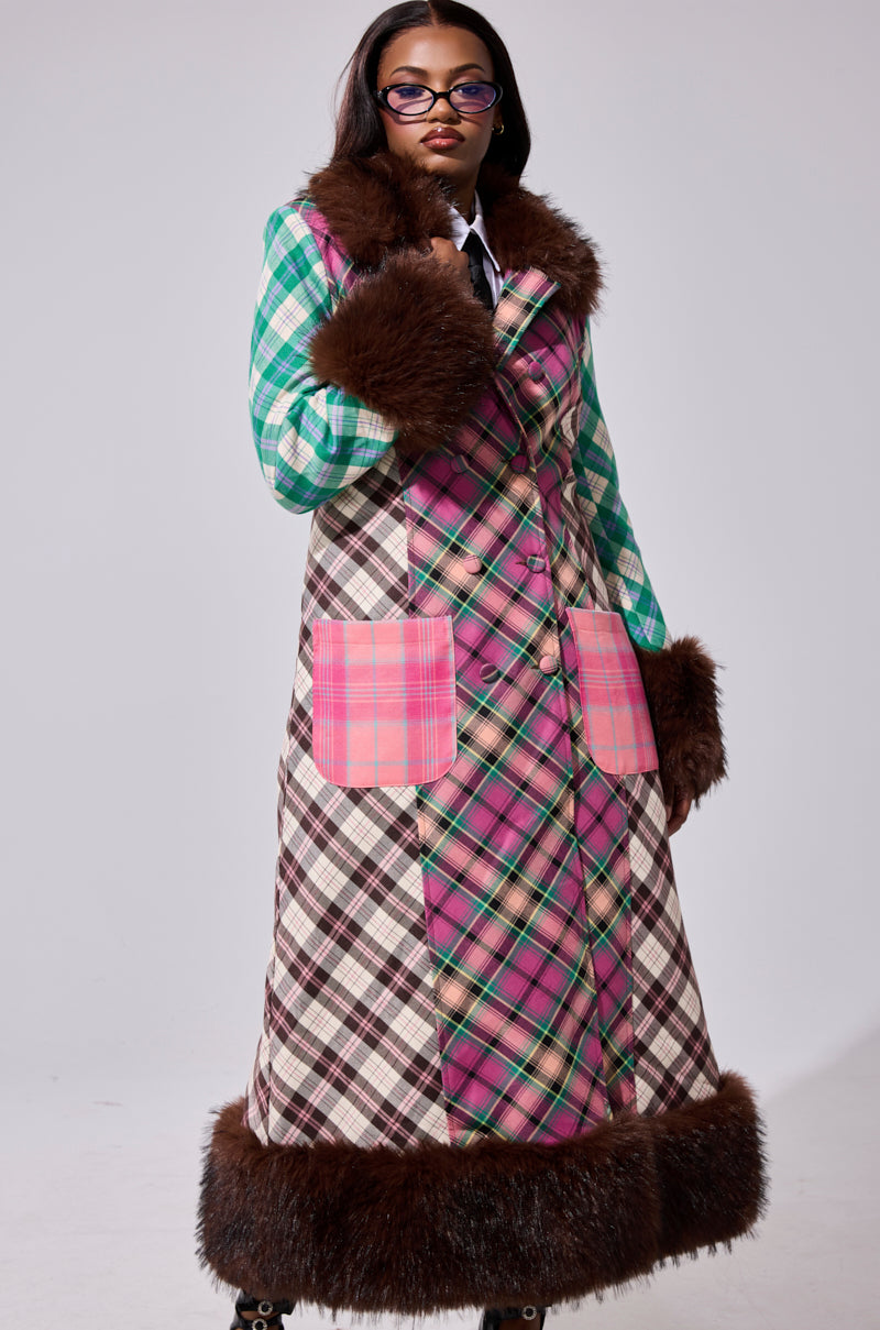A woman in glasses and straight hair stands against a gray background, wearing the SWEET THING PATCHWORK TRENCH—a long coat with pink plaid panels, pink patch pockets, and wide brown faux fur trim on the collar, cuffs, and hem.
