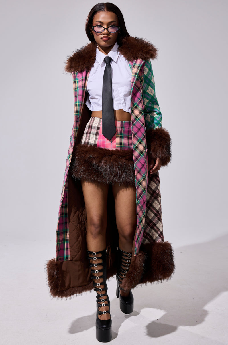 A woman wears round glasses, a white cropped shirt with a black tie, a pink and green plaid mini skirt, the SWEET THING PATCHWORK TRENCH with brown faux fur trim, and black grommet platform boots.