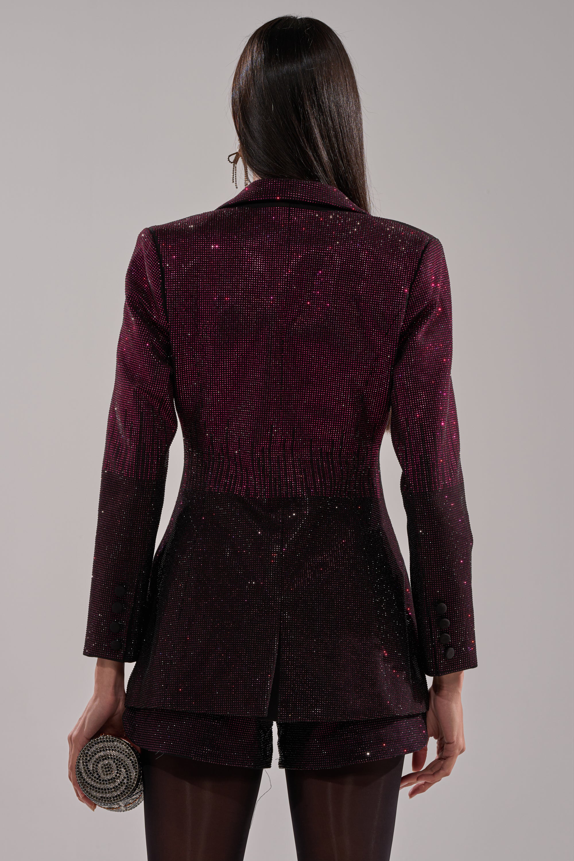 A person with long, straight hair faces away in a dark RAISE A GLASS RHINESTONE BLAZER and matching shorts, holding a small round clutch. The background is plain gray.