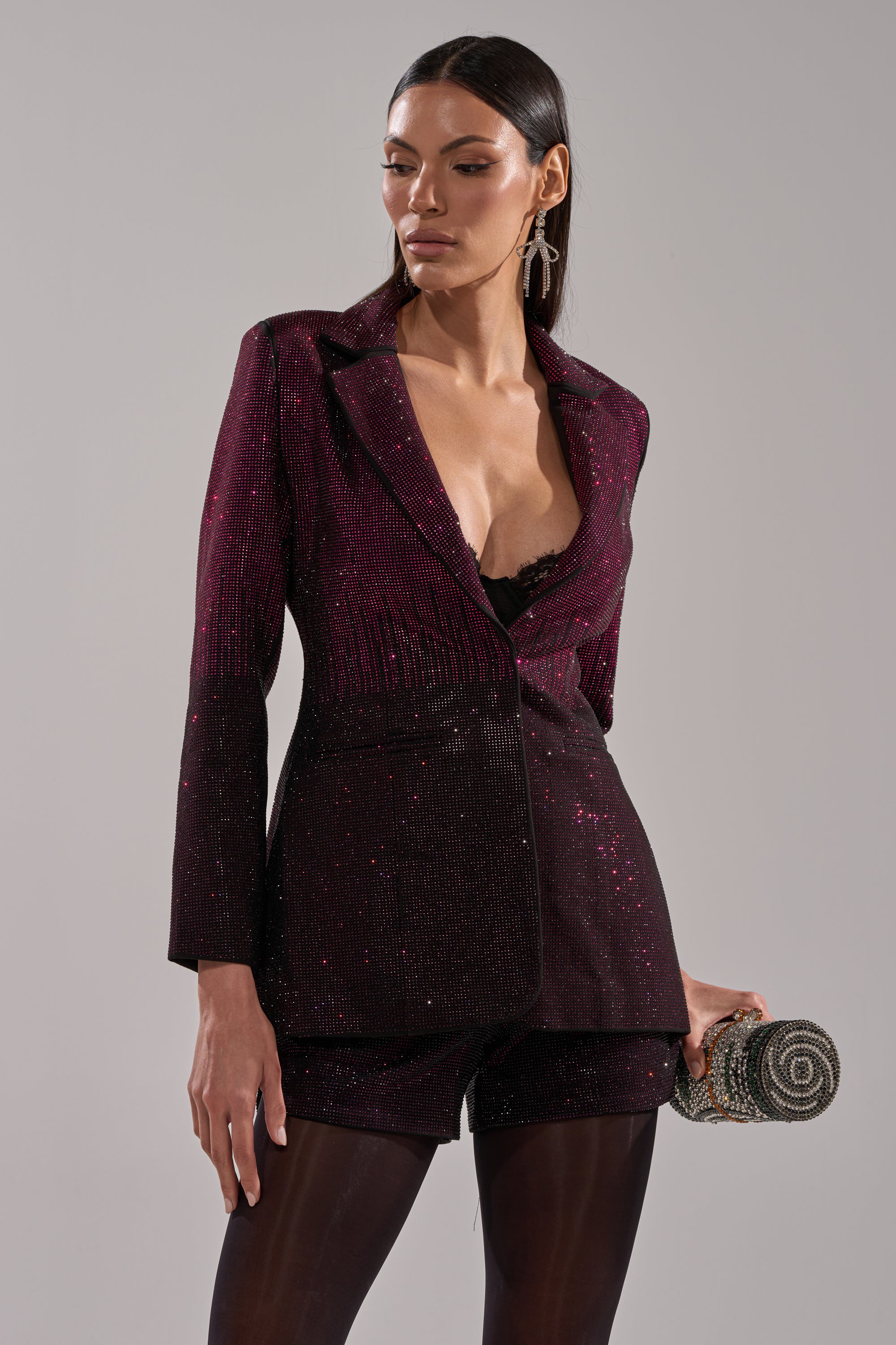 A woman wears the RAISE A GLASS RHINESTONE BLAZER in deep burgundy with matching shorts, a black lace top, sheer tights, a small glittery clutch, drop earrings, and long dark hair for a chic tailored look.