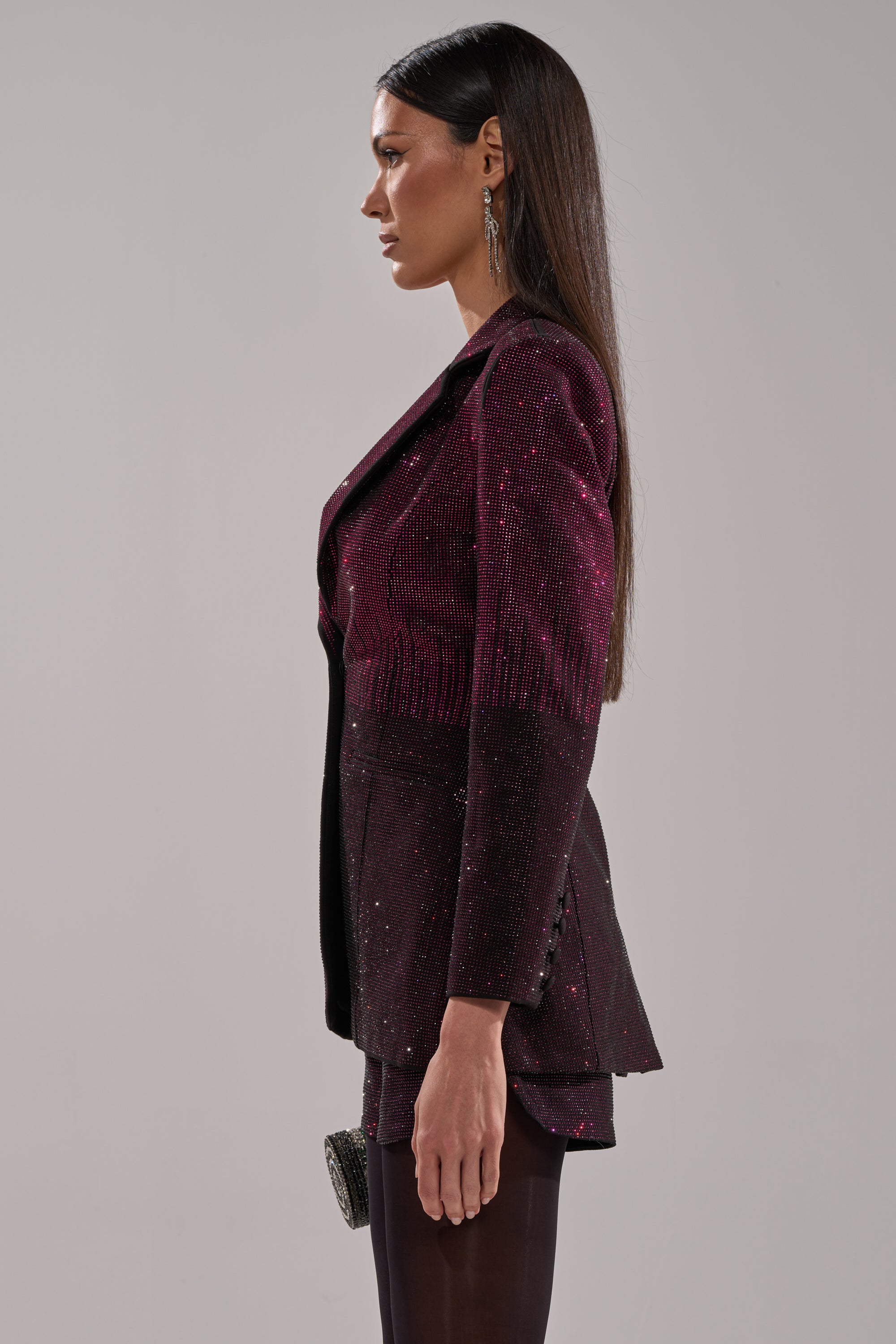 A woman with straight brown hair stands in profile in the AKIRA Label’s RAISE A GLASS RHINESTONE BLAZER and matching skirt, paired with black tights and statement earrings, against a plain gray background.