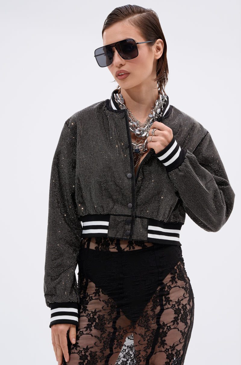 A woman poses confidently against a plain white background wearing the SPOTLIGHT RHINESTONE BOMBER, oversized sunglasses, layered chain necklaces, and sheer black lace pants.