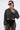 A woman poses confidently against a plain white background wearing the SPOTLIGHT RHINESTONE BOMBER, oversized sunglasses, layered chain necklaces, and sheer black lace pants.