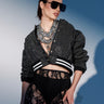 A woman wearing dark sunglasses, a layered necklace, black lace high-waisted pants, and the SPOTLIGHT RHINESTONE BOMBER with striped cuffs poses against a blue and white background.
