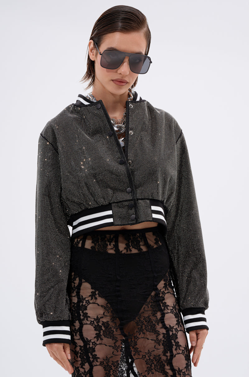 A woman with slicked-back hair wears large sunglasses, a chunky necklace, and the SPOTLIGHT RHINESTONE BOMBER with striped cuffs and hem over high-waisted black lace pants against a plain white background.