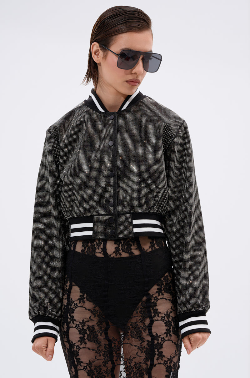 A woman wears the SPOTLIGHT RHINESTONE BOMBER with black and white striped cuffs and waistband, paired with oversized sunglasses and sheer black lace pants over black underwear, standing against a plain light background.
