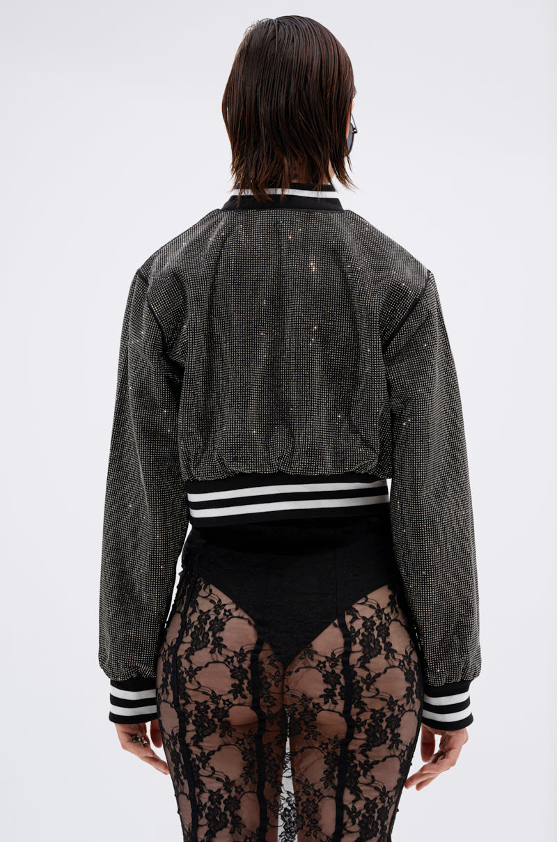 A person with wet, chin-length hair is seen from behind in a SPOTLIGHT RHINESTONE BOMBER with stripes and high-waisted, black lace pants featuring sheer panels. The background is plain white.