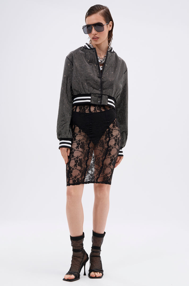 A woman in the SPOTLIGHT RHINESTONE BOMBER, sunglasses, a sheer black lace skirt over high-waisted black underwear, and sheer black heeled socks stands against a plain white background.