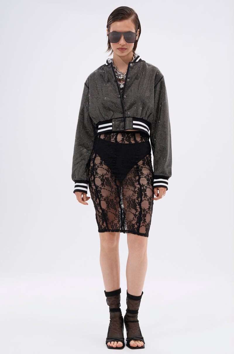 A person poses against a plain background wearing the SPOTLIGHT RHINESTONE BOMBER, a sheer black lace skirt layered over black undergarments, black mesh socks with open-toe heels, layered necklaces, and oversized sunglasses.
