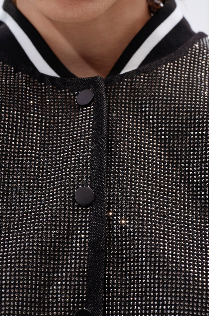 Close-up of the SPOTLIGHT RHINESTONE BOMBER, featuring a varsity-inspired design with sparkling rhinestones, black snap buttons, and a black-and-white striped collar. Only the model’s lower face and neck are visible.