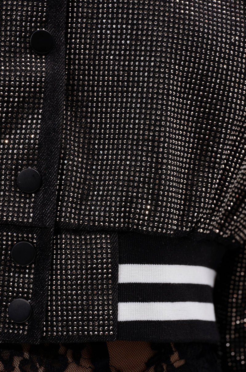 Close-up of the SPOTLIGHT RHINESTONE BOMBER, showcasing shiny rhinestones on black fabric, black snap buttons, and a varsity-style ribbed hem with two white stripes.