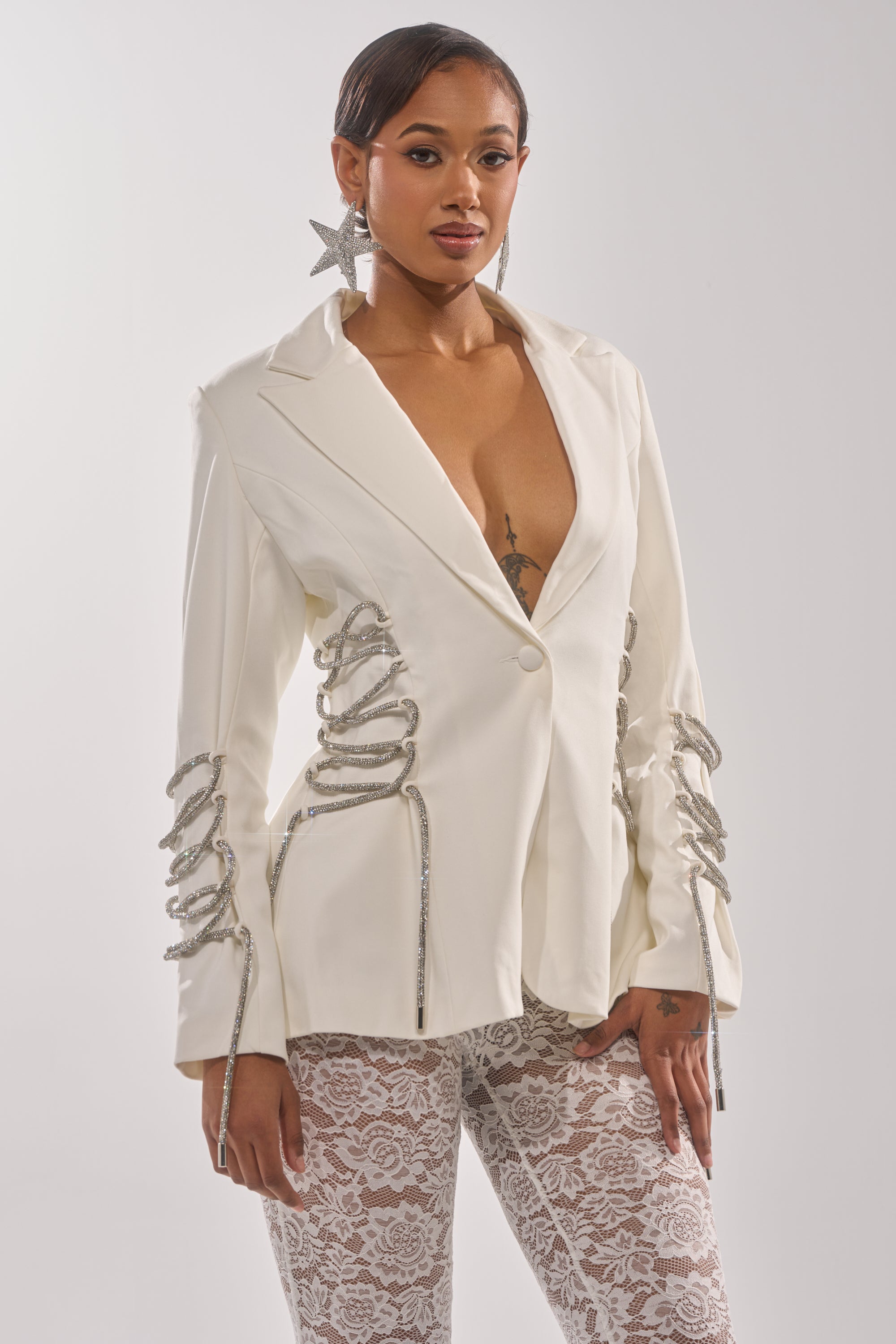 A woman wears the PRETTY PLEASE LACE UP BLAZER, a deep V-neck structured white blazer with silver rope details, styled with star-shaped earrings and white lace pants against a plain light background. 0