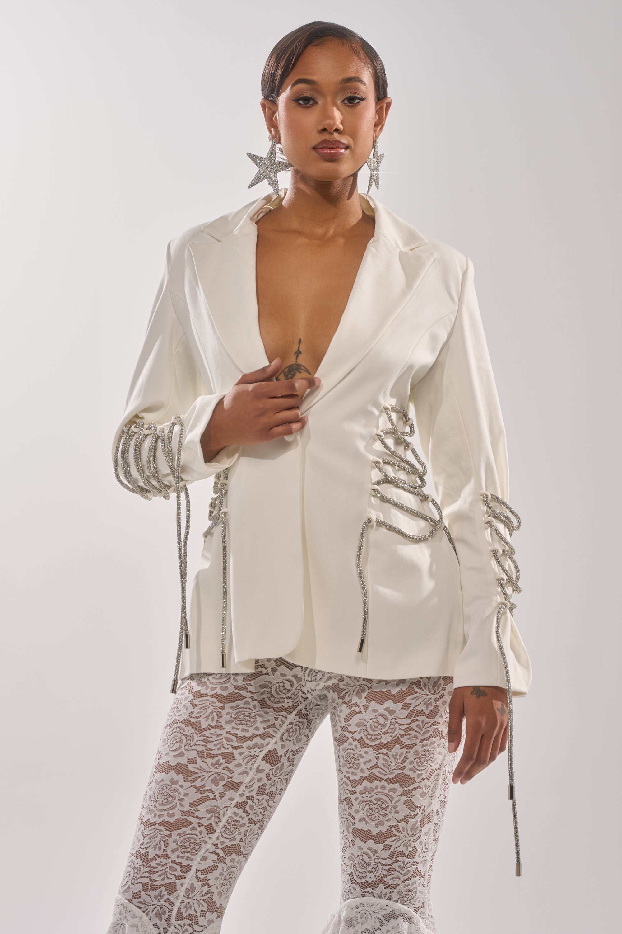 A woman poses confidently in the PRETTY PLEASE LACE UP BLAZER, a white structured piece with a deep neckline and silver rope sleeve details, paired with white lace pants and large star-shaped earrings. Her hair is sleek and styled. 1