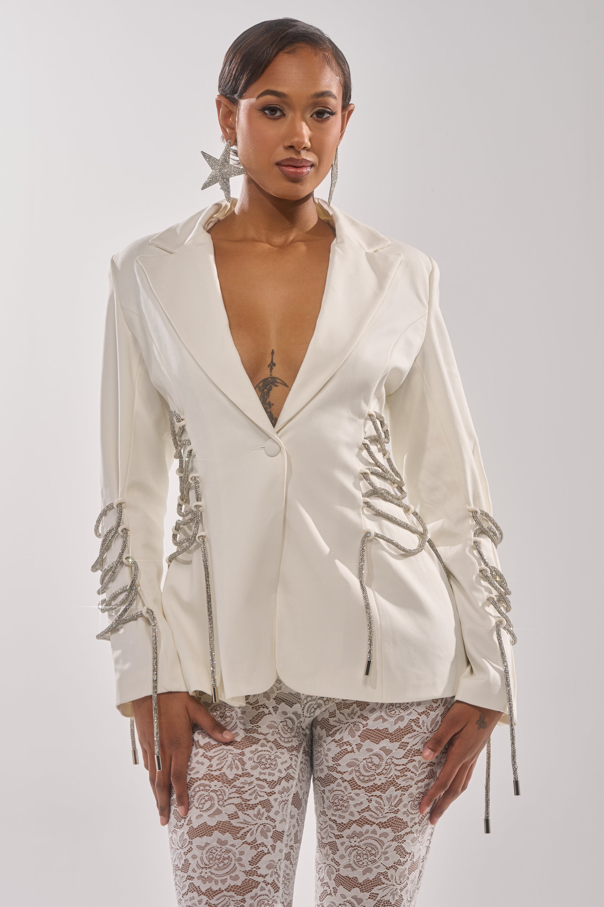 A woman models the PRETTY PLEASE LACE UP BLAZER, a structured white blazer with silver rope lace-up details on the sleeves and sides, styled with white lace pants. She accessorizes with star-shaped earrings and sports a chest tattoo. 2