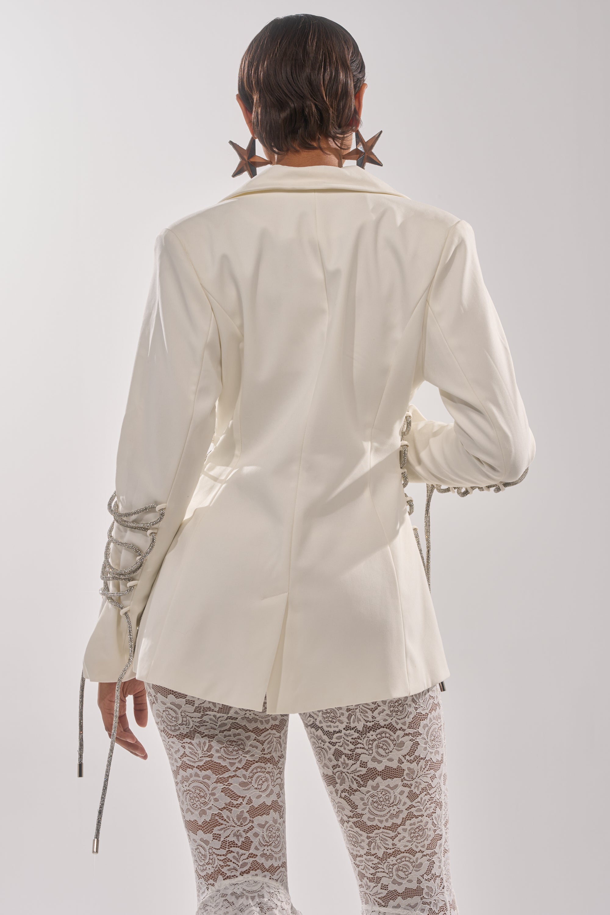 A person faces away, wearing the PRETTY PLEASE LACE UP BLAZER in white with decorative sleeve ties, paired with large star earrings and sheer white lace pants against a plain background. 5