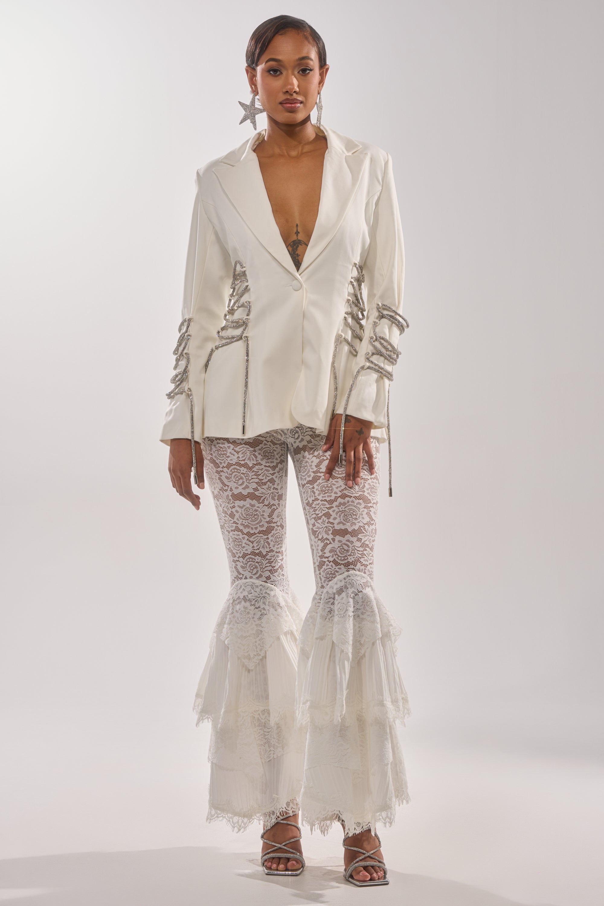 A woman wears the PRETTY PLEASE LACE UP BLAZER, featuring a structured white silhouette with silver chain details, paired with white lace flared pants, statement star earrings, and silver heels against a plain light backdrop. 6