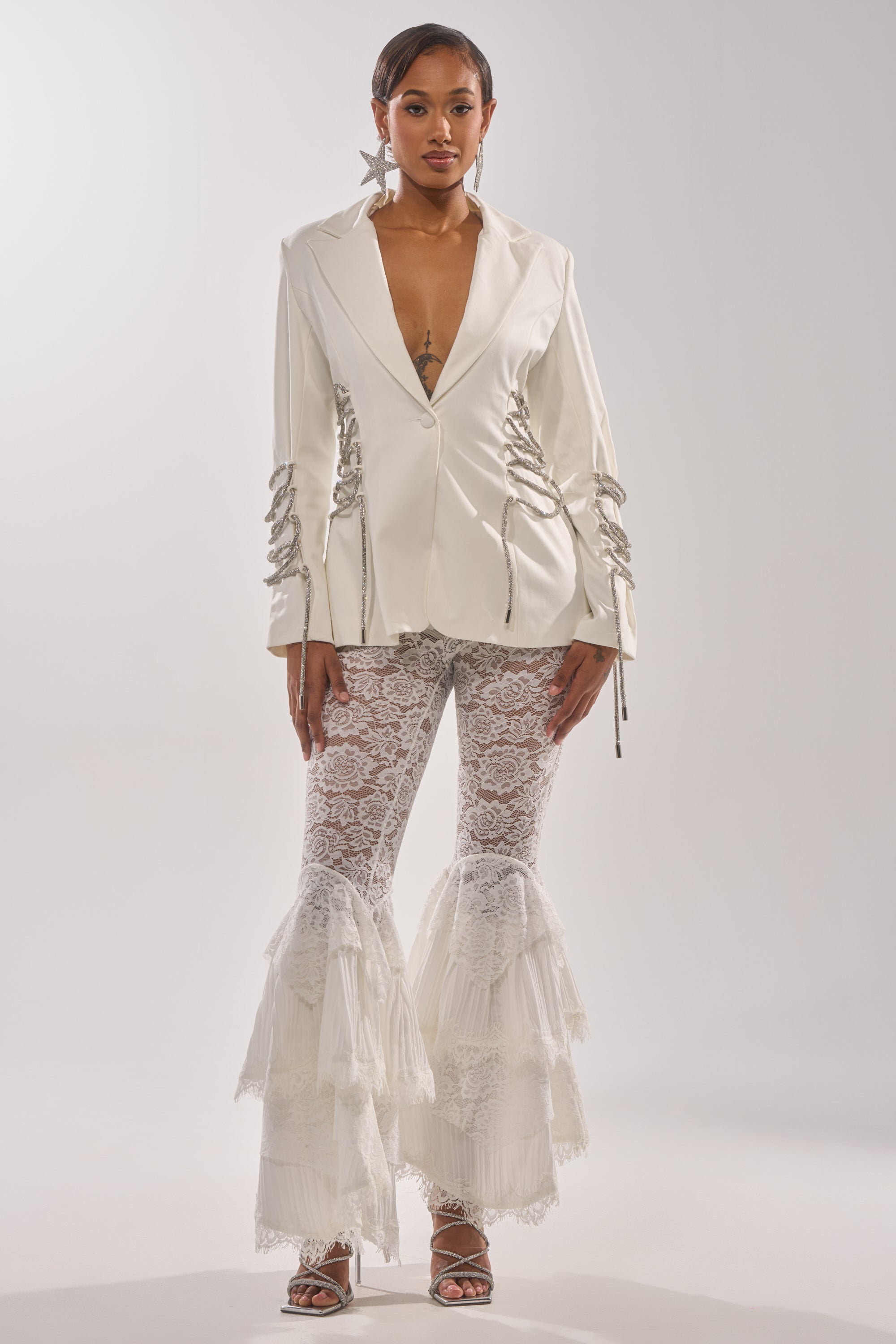 A woman wears the PRETTY PLEASE LACE UP BLAZER in white, featuring silver lace-up details, over sheer lace flared pants with ruffles and strappy heels, standing against a light background. 7
