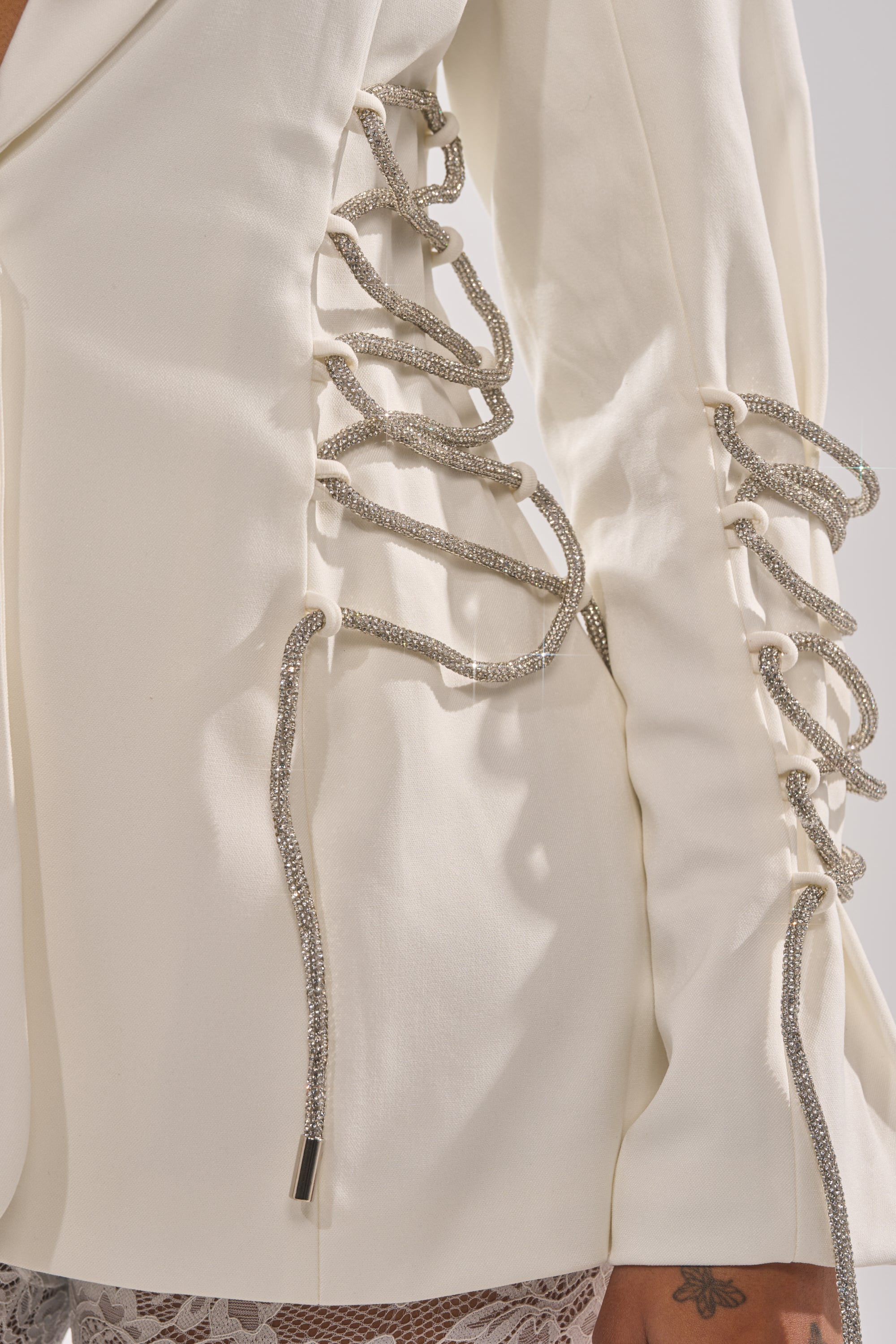 A close-up of the PRETTY PLEASE LACE UP BLAZER’s sleeve, showing decorative silver rope lacing with metallic tips over lace fabric, highlighting its structured silhouette. 8