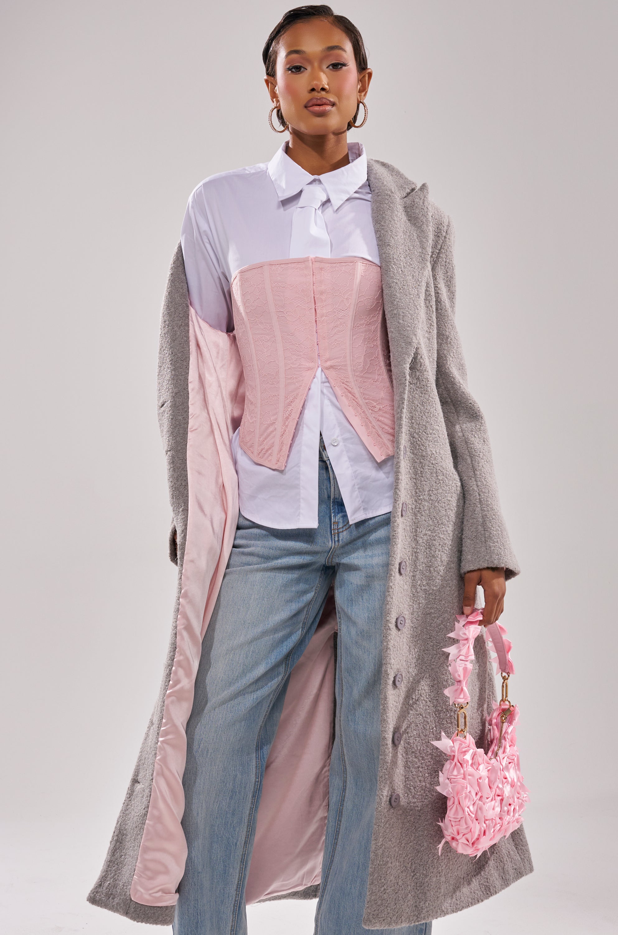 A woman styles the MAKE IT LAST BOUCLE TRENCH—a long gray coat with blush pink lining—over a white button-up, light pink corset, light blue jeans, and carries a pink ruffled handbag.