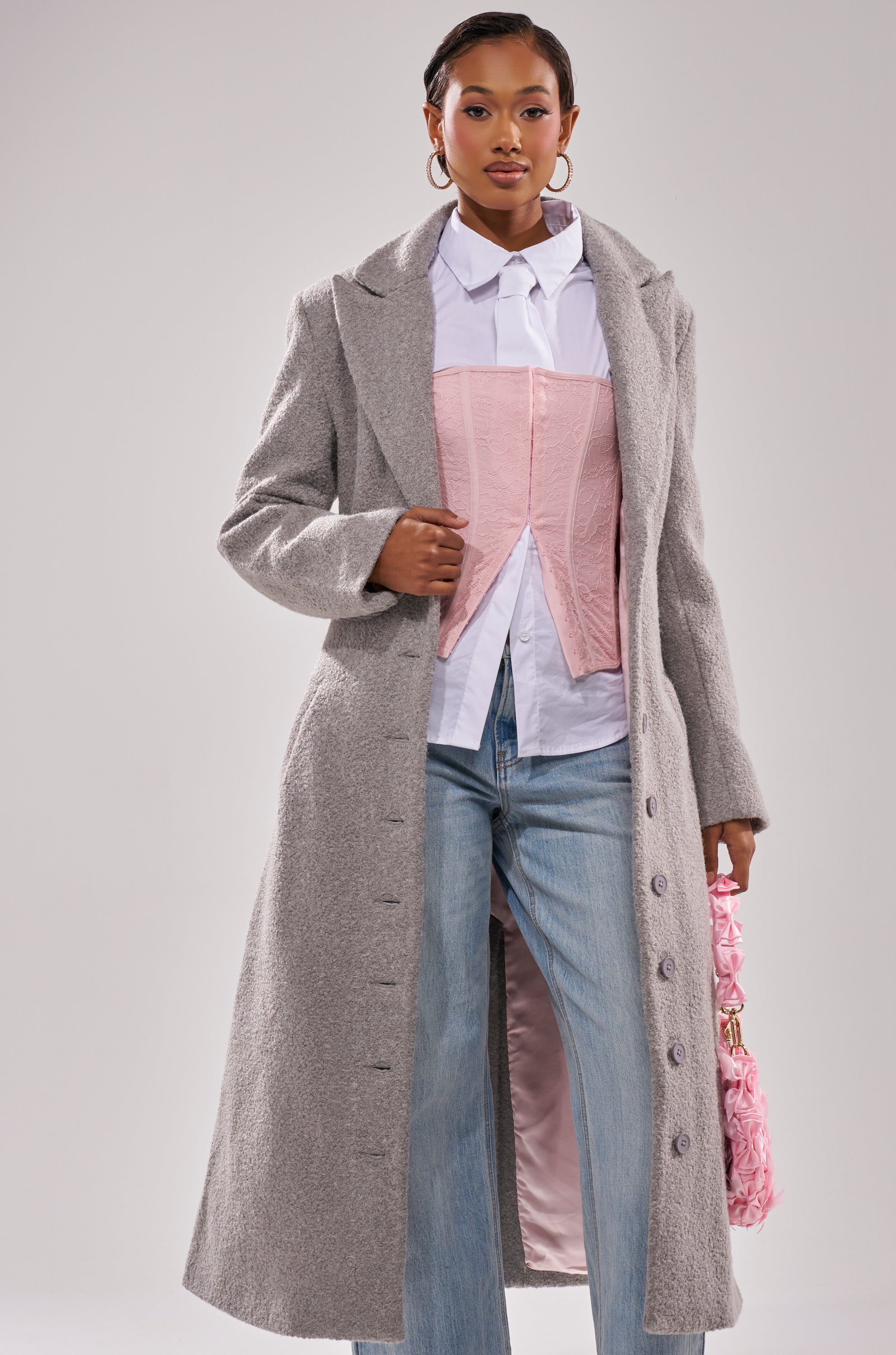 A woman stands against a plain background wearing the MAKE IT LAST BOUCLE TRENCH—a long gray coat—over a white button-up shirt and pink corset, paired with light blue jeans and a pink textured handbag.