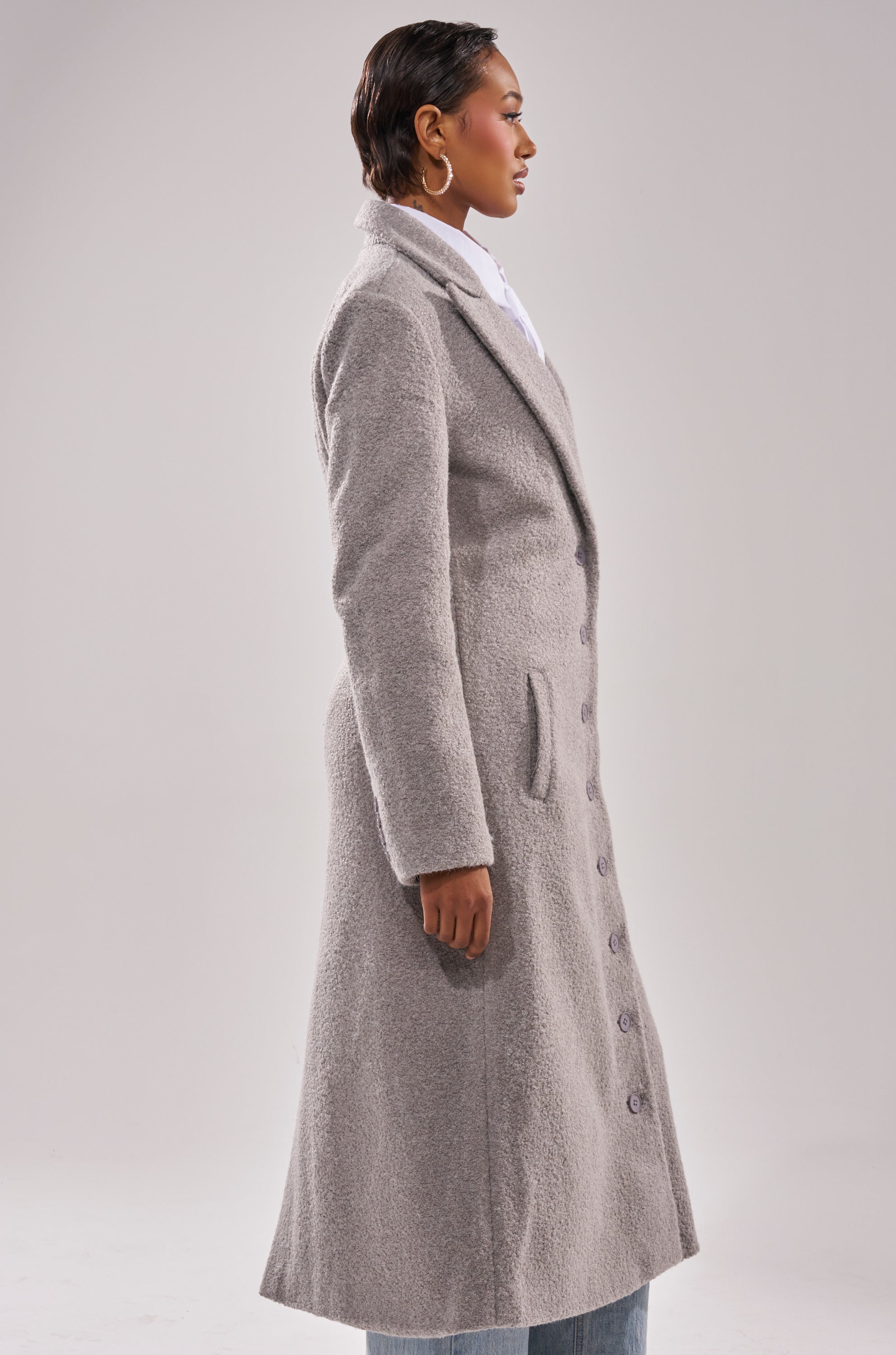 Wearing the MAKE IT LAST BOUCLE TRENCH, a person stands sideways against a plain light background. The textured, light gray trench features front buttons, side pockets, and is styled with light pants.