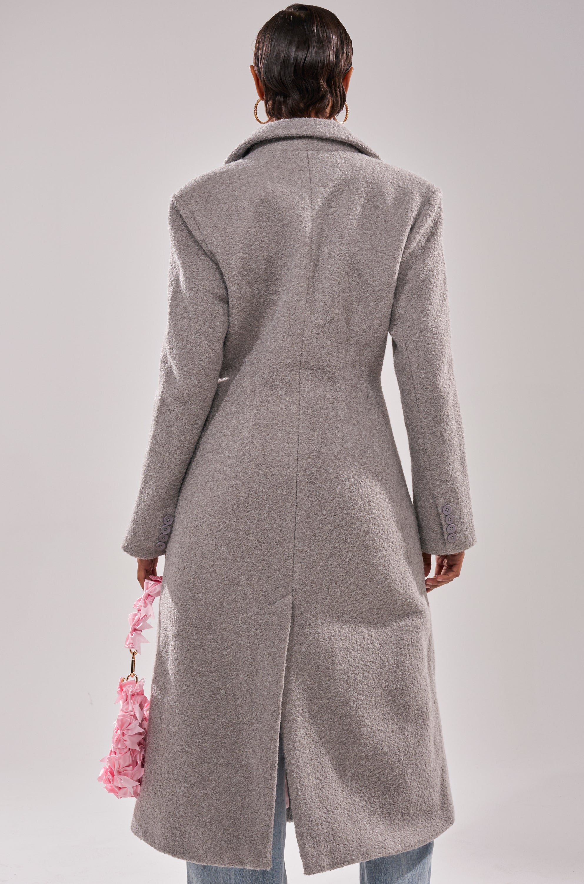Seen from behind, a person in the MAKE IT LAST BOUCLE TRENCH with a longline silhouette and blue jeans carries a pink ruffled handbag.