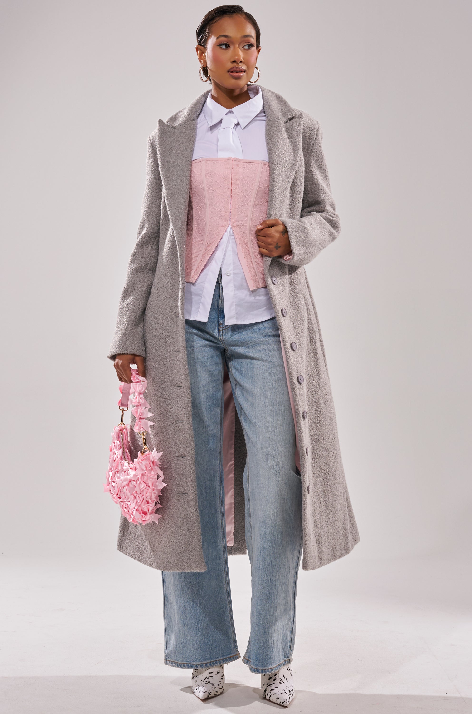 A woman wears the MAKE IT LAST BOUCLE TRENCH over a white shirt and pink corset, paired with light blue wide-leg jeans, white boots, a pink textured handbag, and hoop earrings while posing against a plain background.