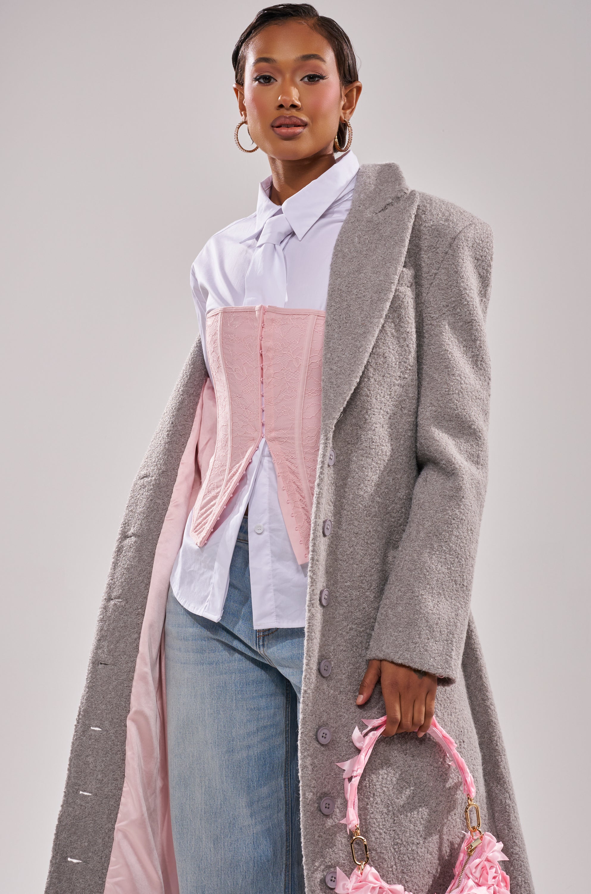A woman styles the MAKE IT LAST BOUCLE TRENCH over a white button-up shirt and light pink corset, paired with light blue jeans. She carries a pink handbag, wears gold hoop earrings, and has neatly styled hair and makeup.