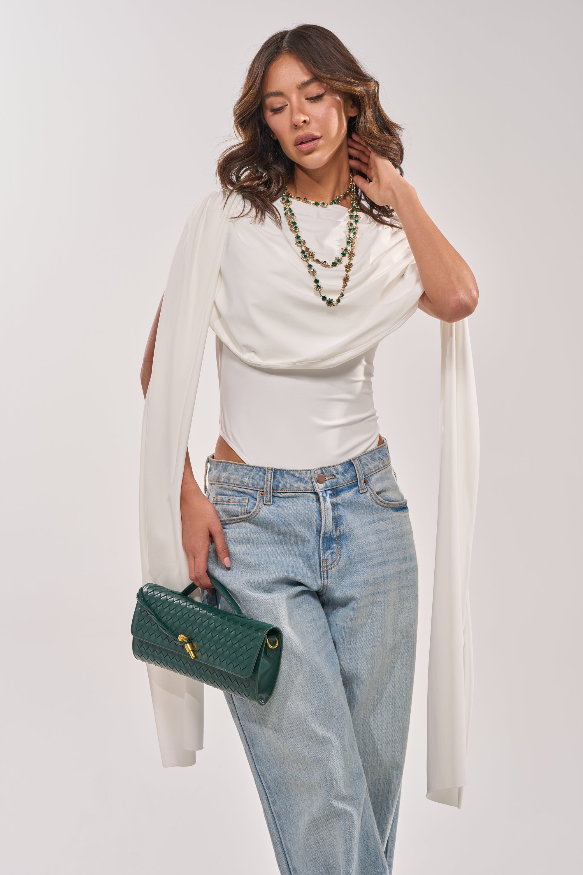 A woman with wavy brown hair stands against a light background wearing the HILLARY SEAMLESS BODYSUIT IN IVORY, light blue jeans, layered green bead necklaces, and holding a green quilted handbag. 0