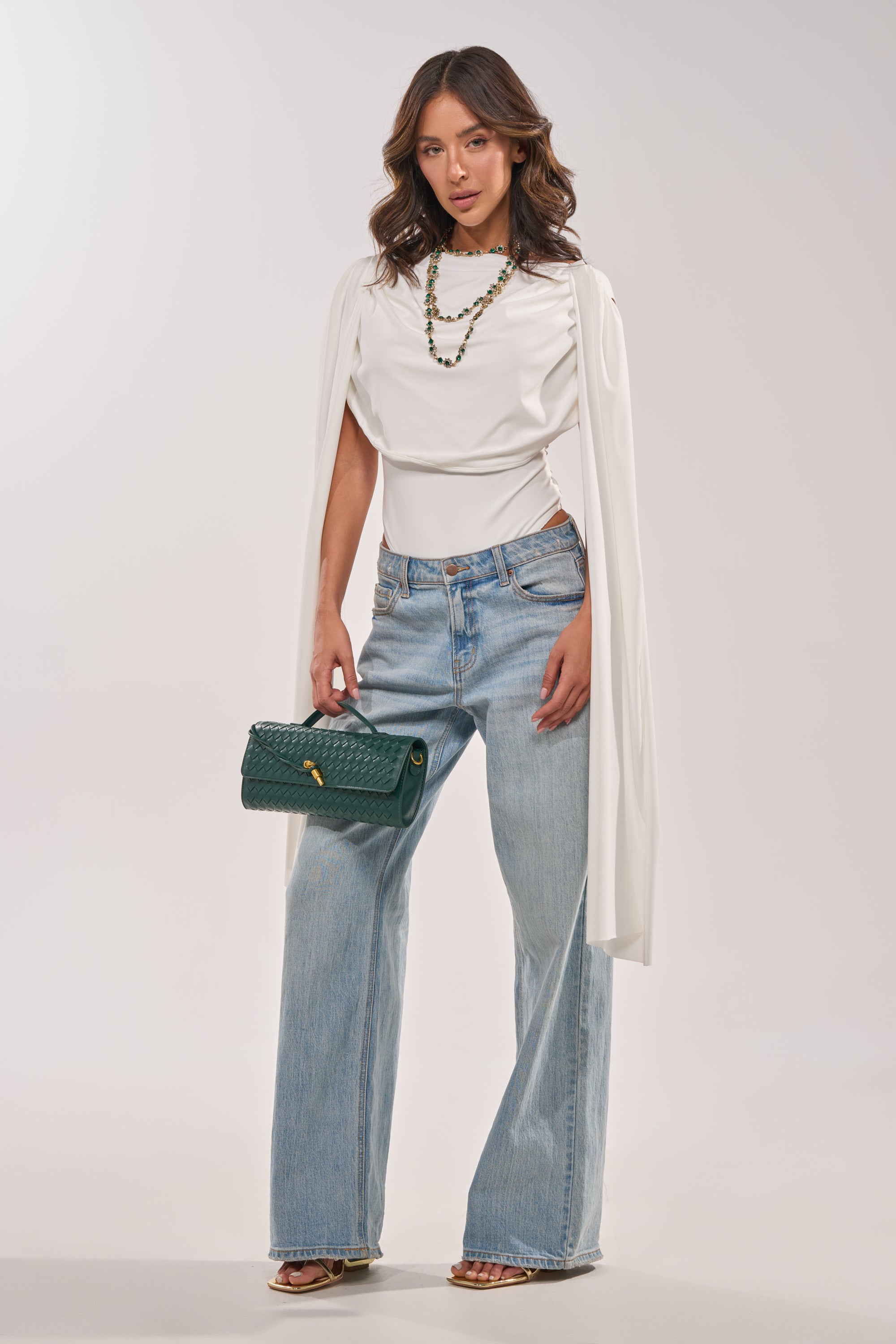 A woman wears the HILLARY SEAMLESS BODYSUIT IN IVORY with wide-leg light blue jeans, heeled sandals, a green clutch, and layered necklaces as she poses against a plain light background. 1