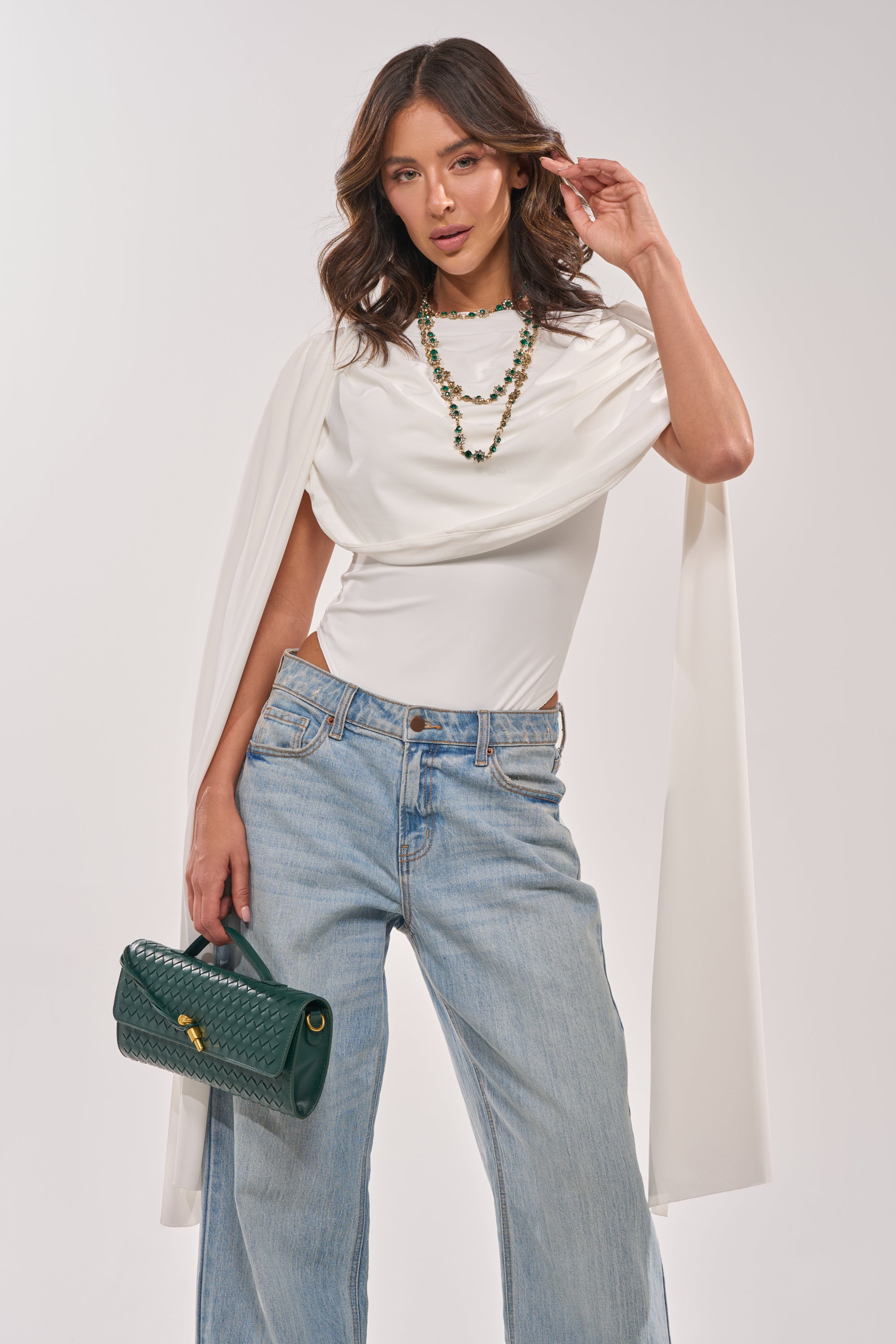A woman with wavy brown hair and a confident look wears the HILLARY SEAMLESS BODYSUIT IN IVORY, light blue wide-leg jeans, a green beaded necklace, and carries a green textured handbag. 2
