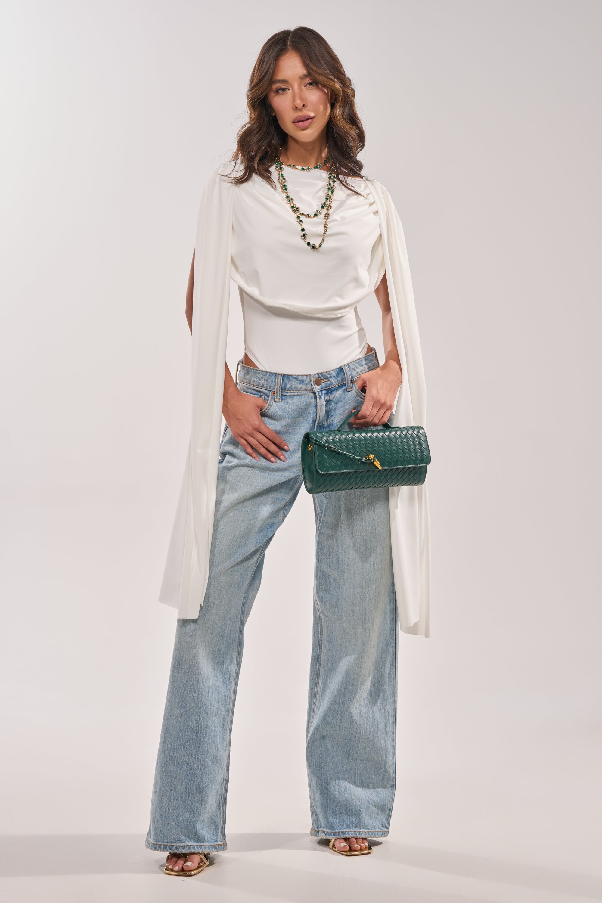 A woman stands confidently in the Hillary Seamless Bodysuit in Ivory, paired with light blue wide-leg jeans, gold sandals, layered necklaces, and a green textured handbag against a plain light background. 4