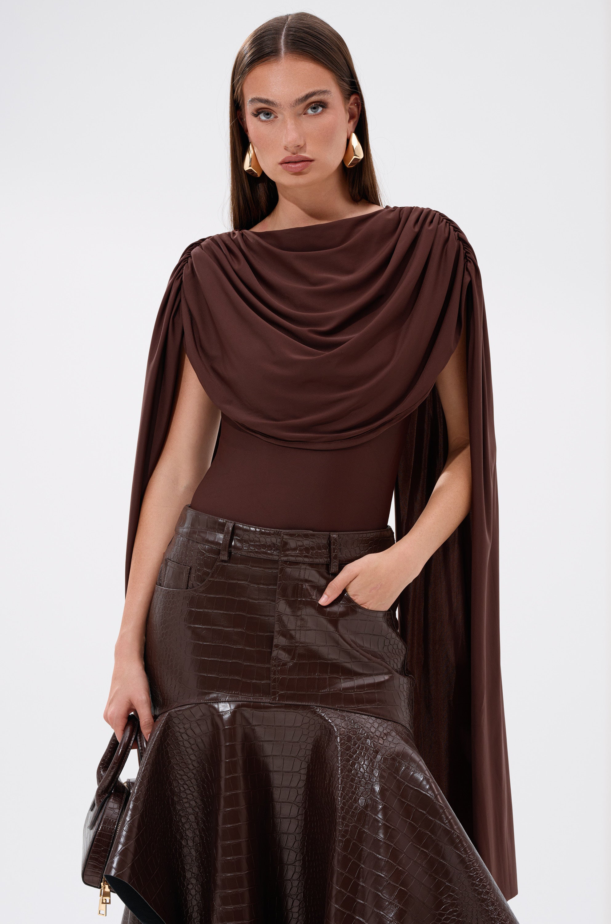 A woman wears the HILLARY SEAMLESS BODYSUIT with draped cape-like sleeves, paired with a shiny brown crocodile-patterned skirt and matching purse. She has long brown hair, gold geometric earrings, and stands against a plain white background.