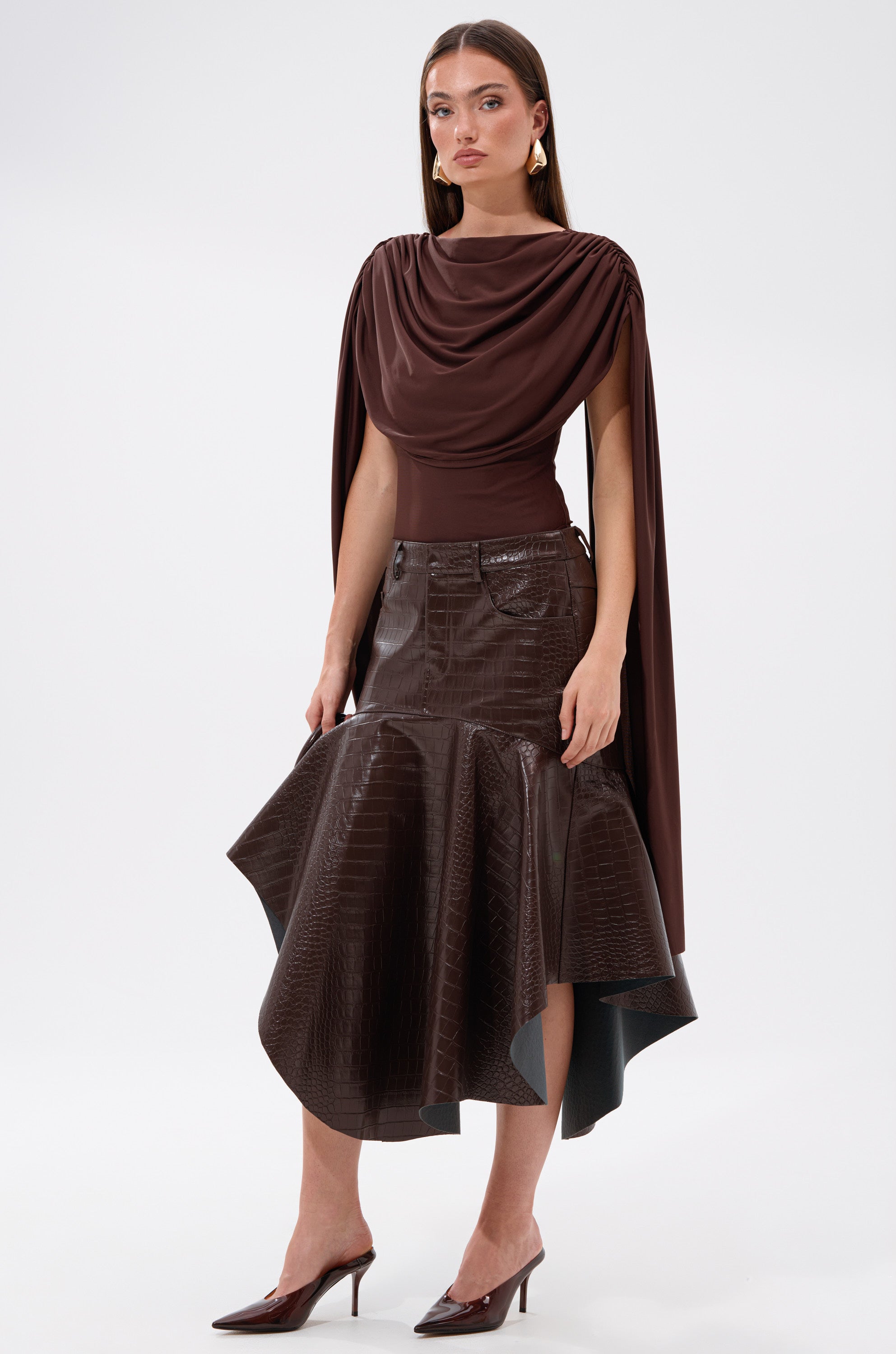 A woman in the HILLARY SEAMLESS BODYSUIT stands against a plain white background, styled with a glossy dark brown asymmetrical skirt and pointed brown heeled mules.