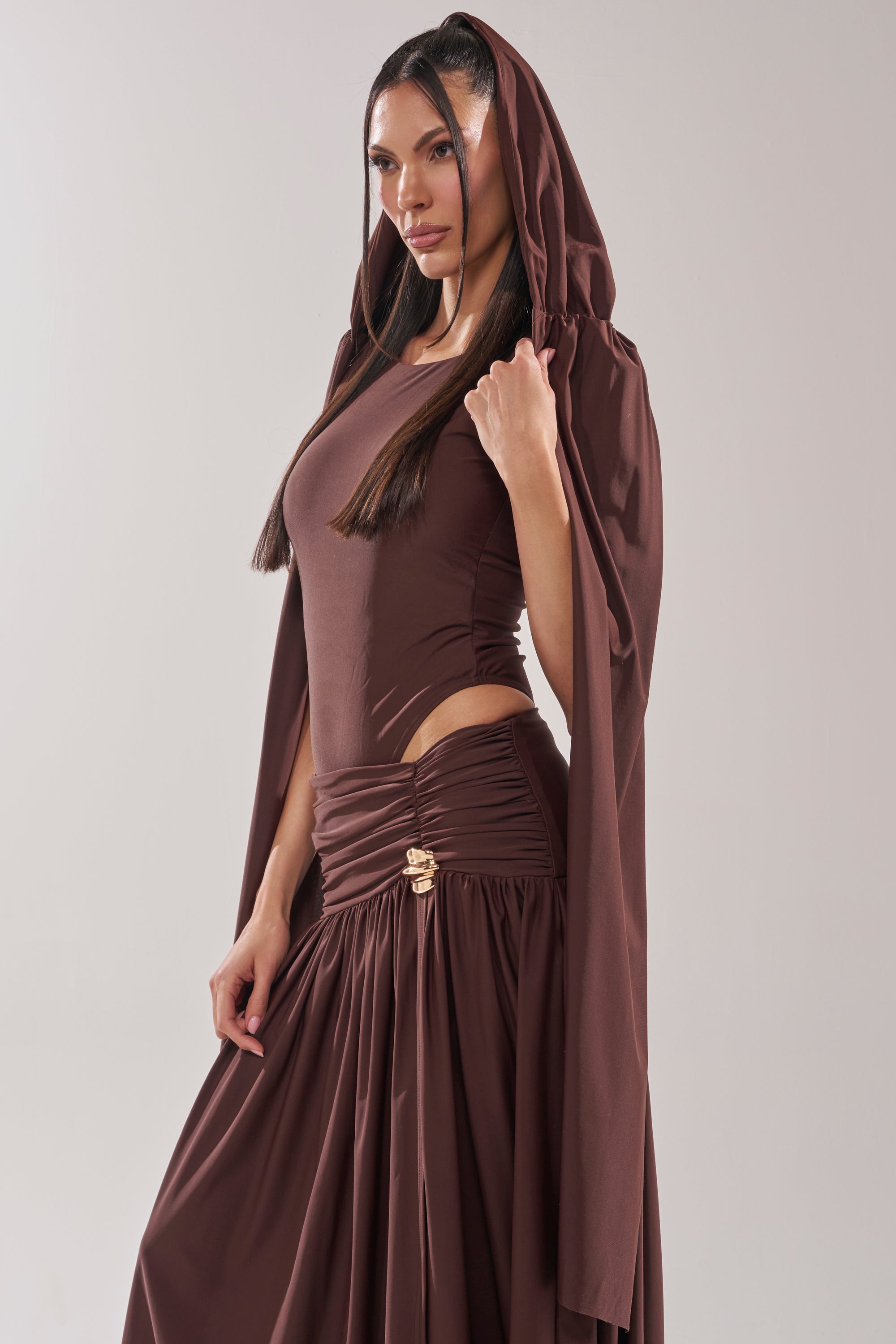 Wearing the HILLARY SEAMLESS BODYSUIT in chocolate brown, a woman lifts her hood to reveal a subtle cape overlay. The fitted bodysuit is paired with a flowing skirt featuring gold accents and gathered waist details as she gazes to the side. 6