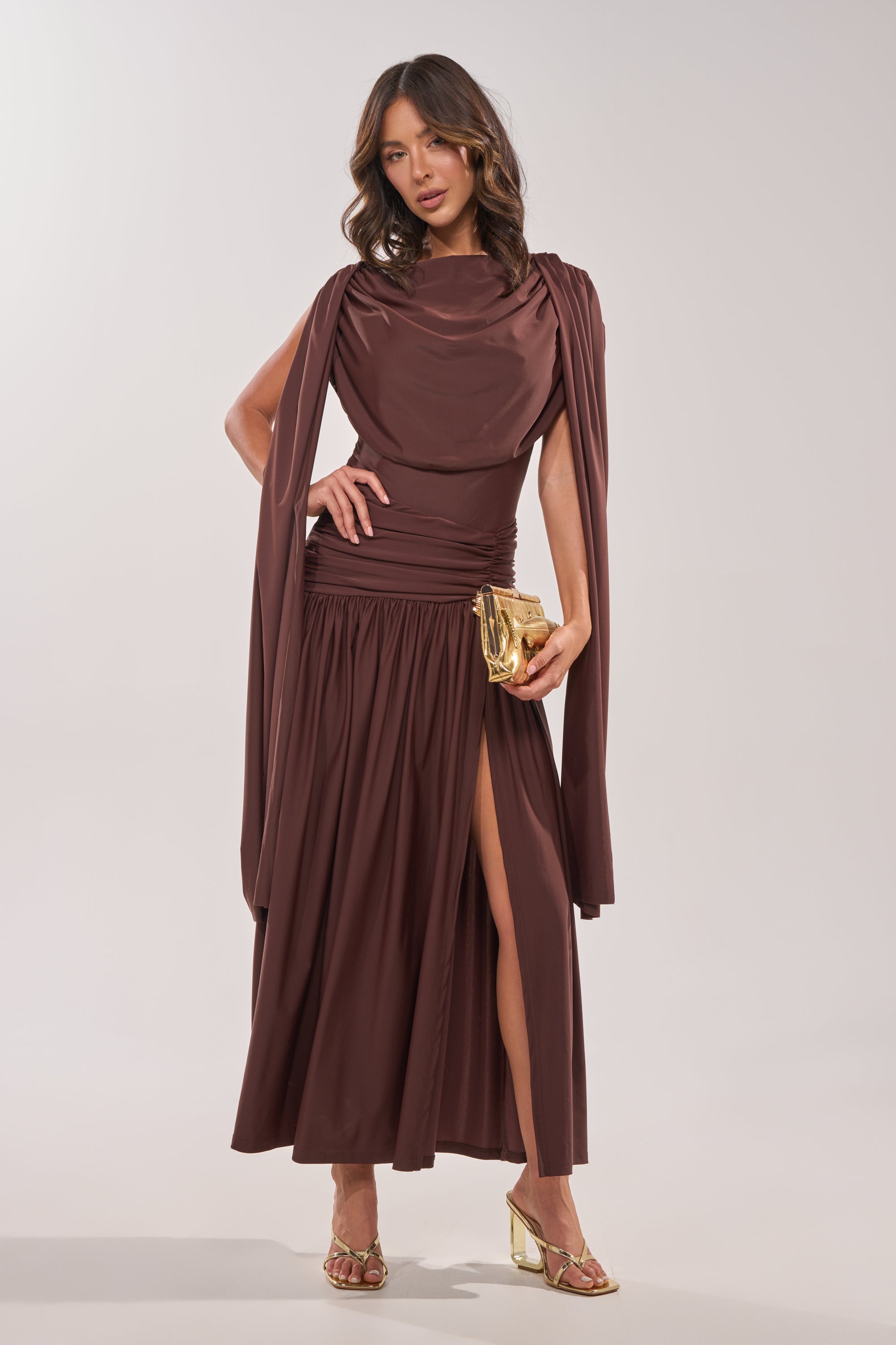 A woman models the HILLARY SEAMLESS BODYSUIT in chocolate brown, featuring a draped design with flowing sleeves and a high neckline. She accessorizes with a gold clutch and heels, set against a simple light background. 0