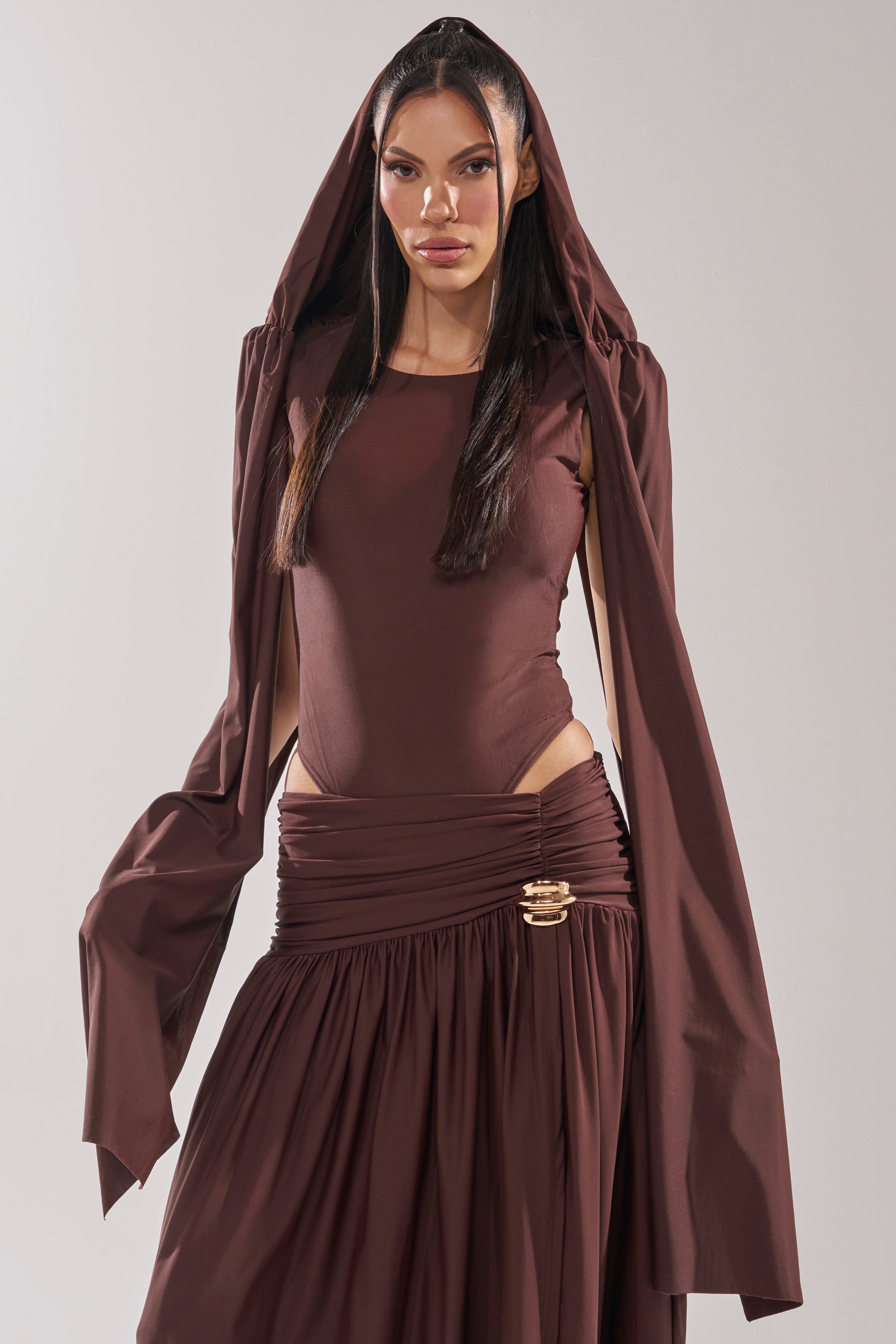 A woman wears the HILLARY SEAMLESS BODYSUIT in chocolate brown, featuring long sleeves, a hood, and a matching skirt with a gold brooch. She stands against a plain background, her elegant look enhanced by a subtle cape overlay. 7