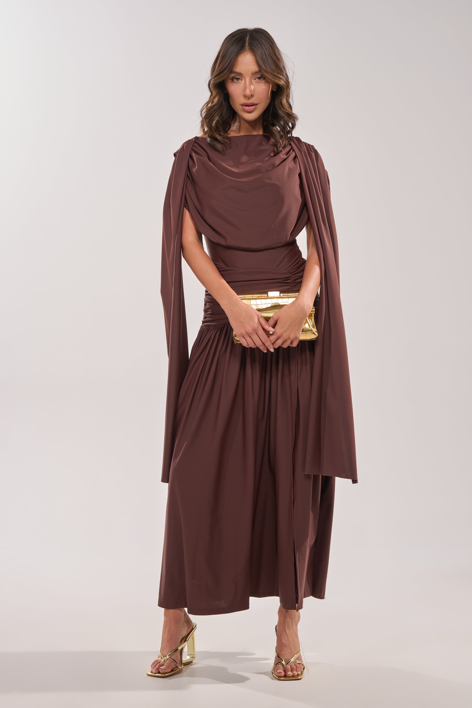 A woman faces forward in the HILLARY SEAMLESS BODYSUIT with cape-like sleeves, holding a gold clutch and wearing gold sandals against a plain, light-colored background. 1