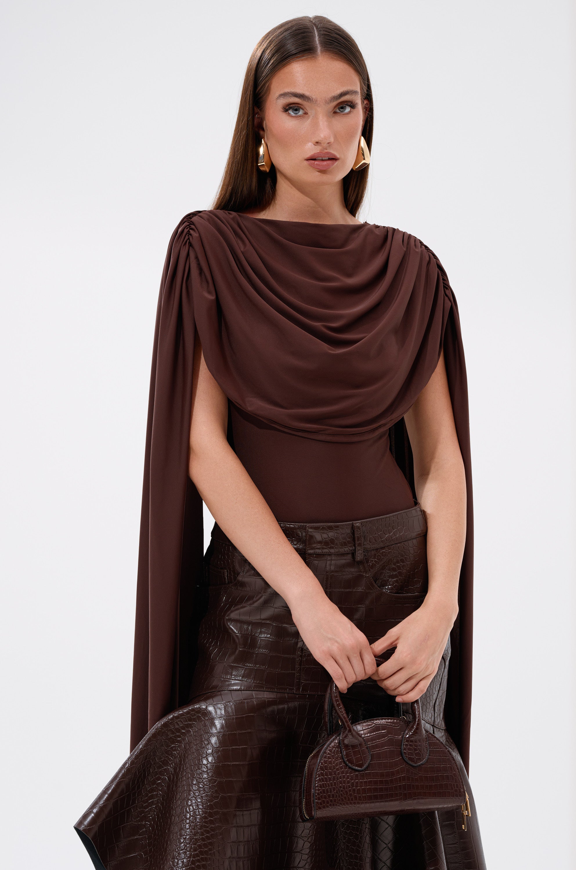 A woman with long brown hair wears the HILLARY SEAMLESS BODYSUIT with dramatic cape sleeves, a textured leather skirt, gold earrings, and holds a small brown purse. She stands against a plain background with a neutral expression.