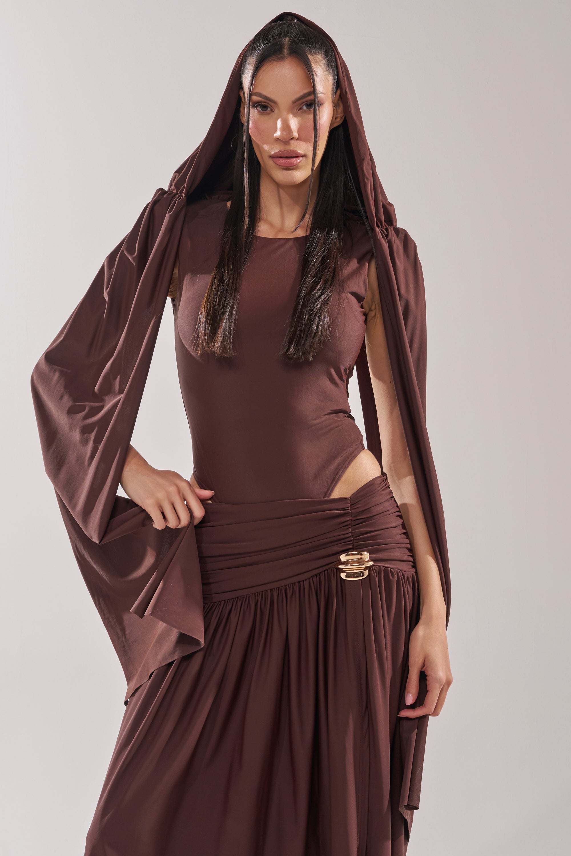 A woman in the HILLARY SEAMLESS BODYSUIT, featuring a chocolate brown hue with a draped skirt and matching hooded wrap, poses confidently against a plain light background, her long straight dark hair framing her neutral expression. 8