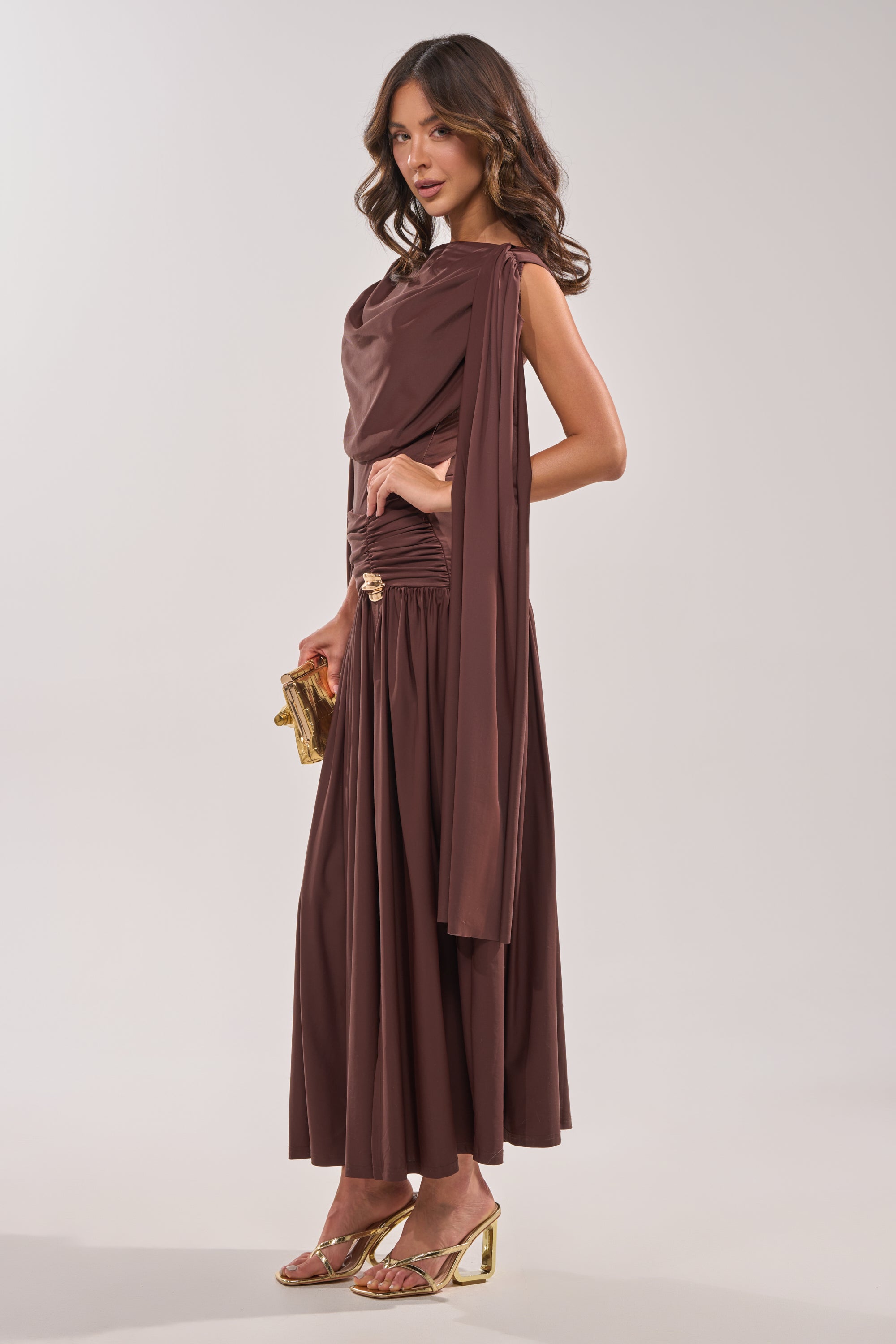 A woman in the HILLARY SEAMLESS BODYSUIT stands sideways, holding a small gold clutch. She wears gold strappy heeled sandals and has loose, wavy brown hair against a plain light background. 3