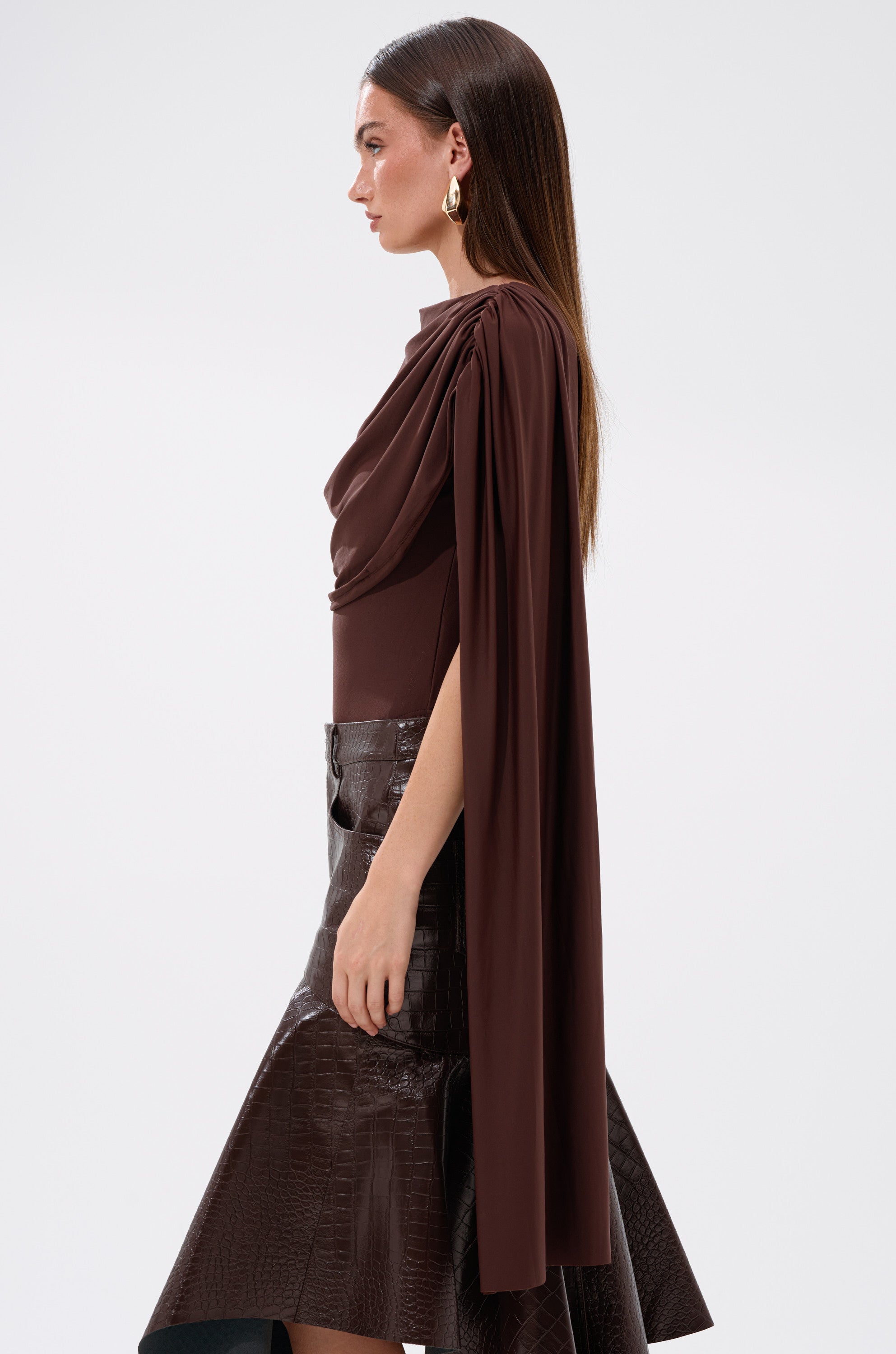 A woman poses in profile wearing the HILLARY SEAMLESS BODYSUIT in chocolate brown with draped, cape-like sleeves, styled with a textured brown skirt and gold hoops. Her straight hair is worn down against a plain, light background.