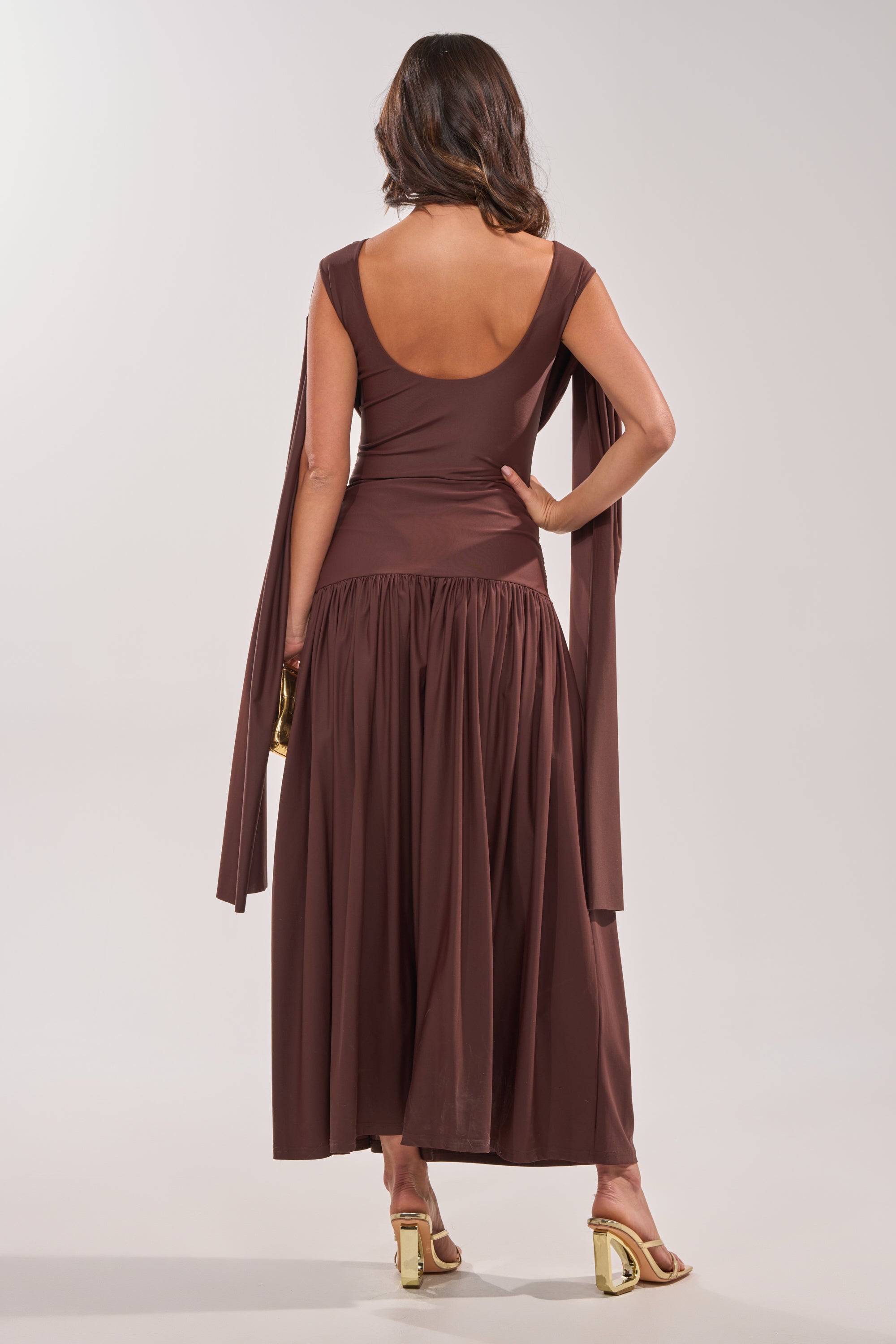 Woman stands with her back to the camera, wearing the HILLARY SEAMLESS BODYSUIT in chocolate brown, featuring an open back and long sleeves. She accessorizes with a small gold clutch and gold heeled sandals. 4