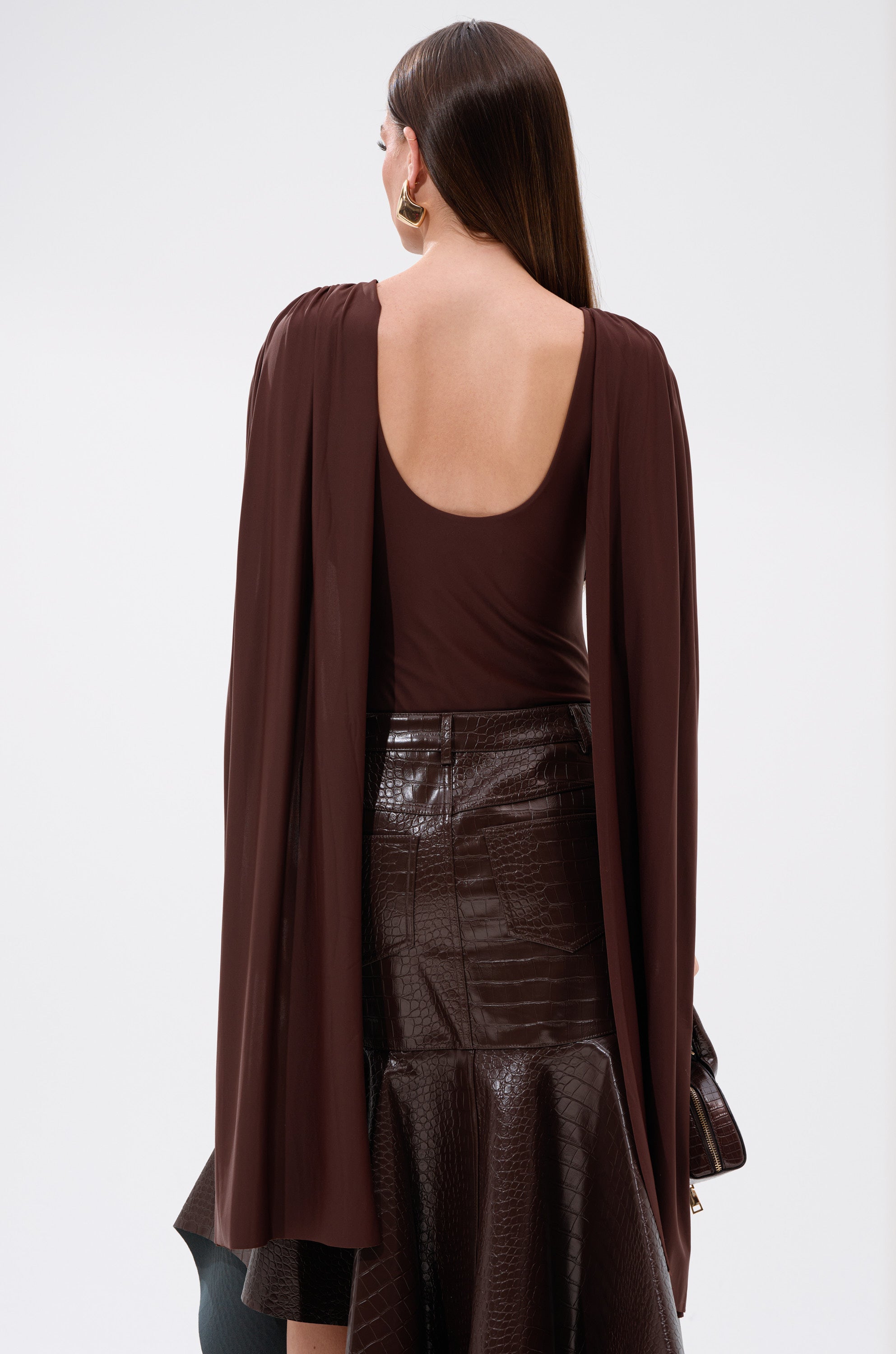 A woman with long brown hair faces away from the camera, wearing the HILLARY SEAMLESS BODYSUIT in chocolate brown with an open back and paired with a high-waisted, dark brown flared skirt against a plain background.