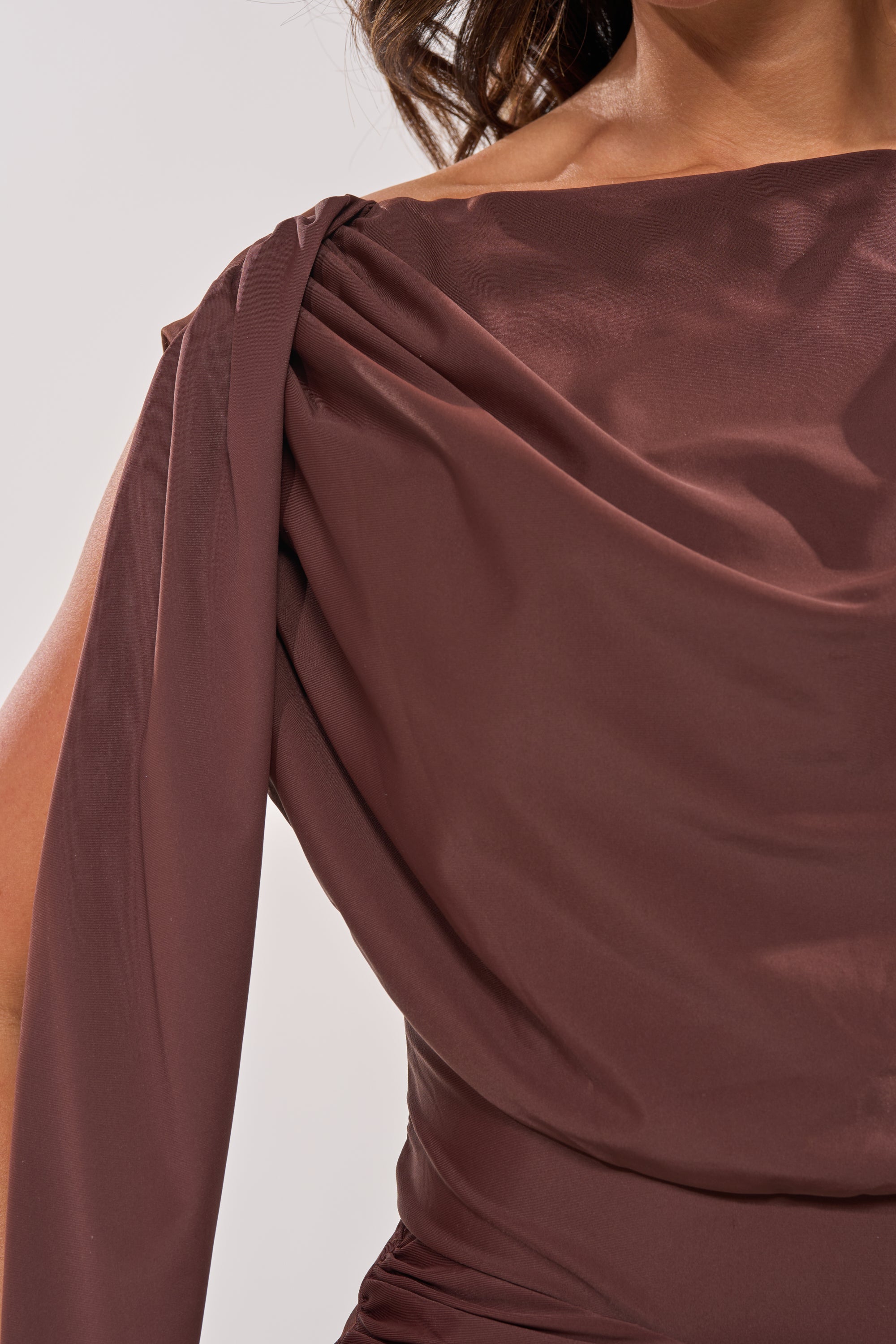 A close-up of a woman in the HILLARY SEAMLESS BODYSUIT, showcasing its silky, off-the-shoulder design with long sleeves and draped fabric. Only part of her upper body and arm is visible against a plain background. 5