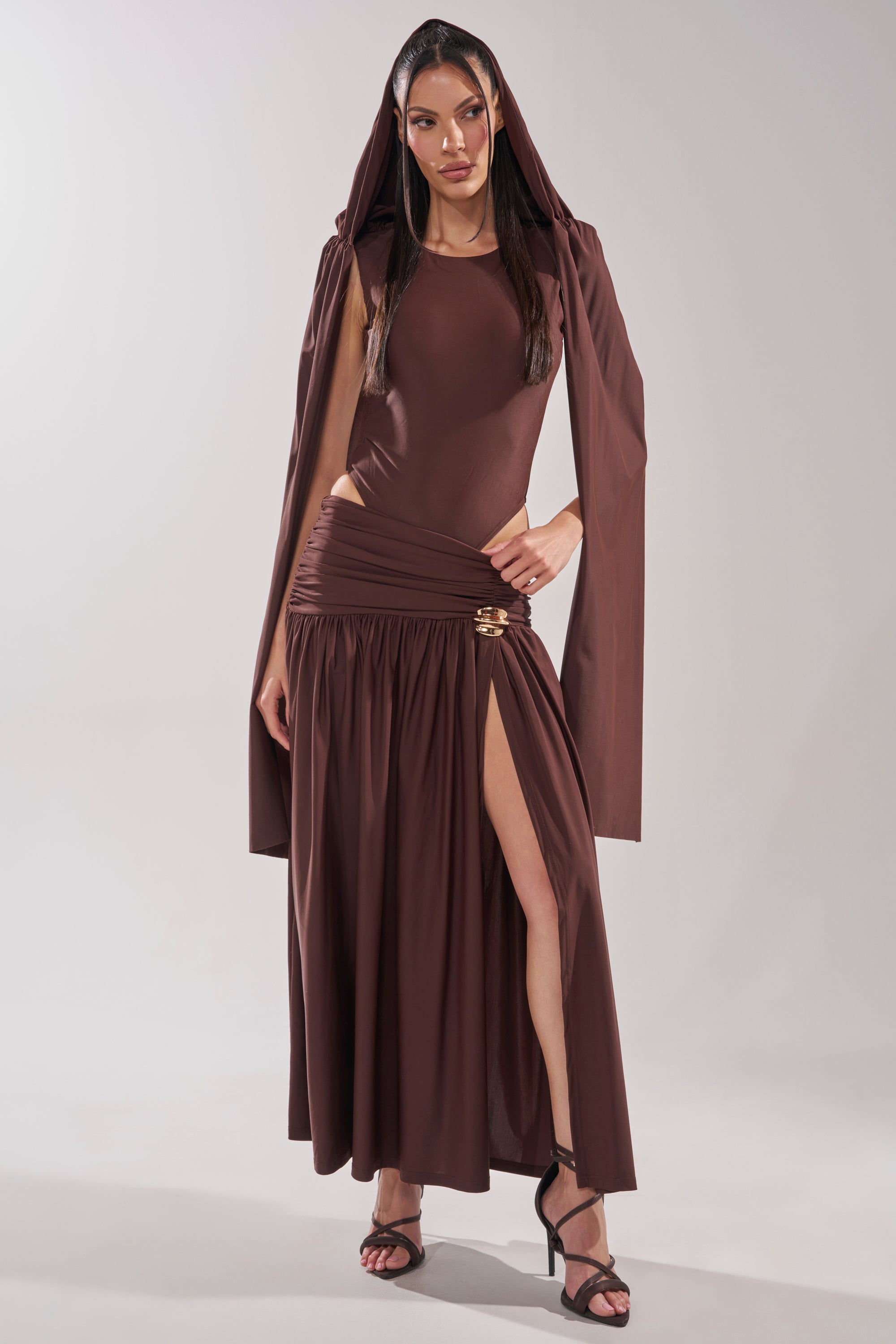 A woman models the HILLARY SEAMLESS BODYSUIT, a stylish brown design with a hooded long-sleeve top, fitted bodice, and high-slit skirt, paired with black strappy heels against a plain light background. 9